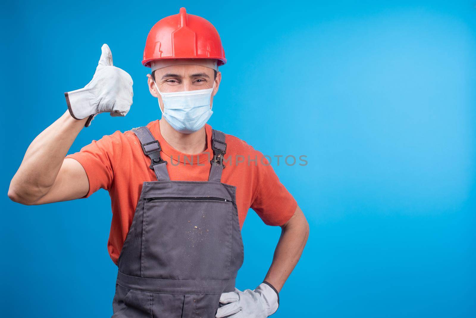 Confident male builder in uniform with gloves, face mask and helmet holding hand on waist, looking at camera and gesturing thumb up while approving work against blue background during epidemic