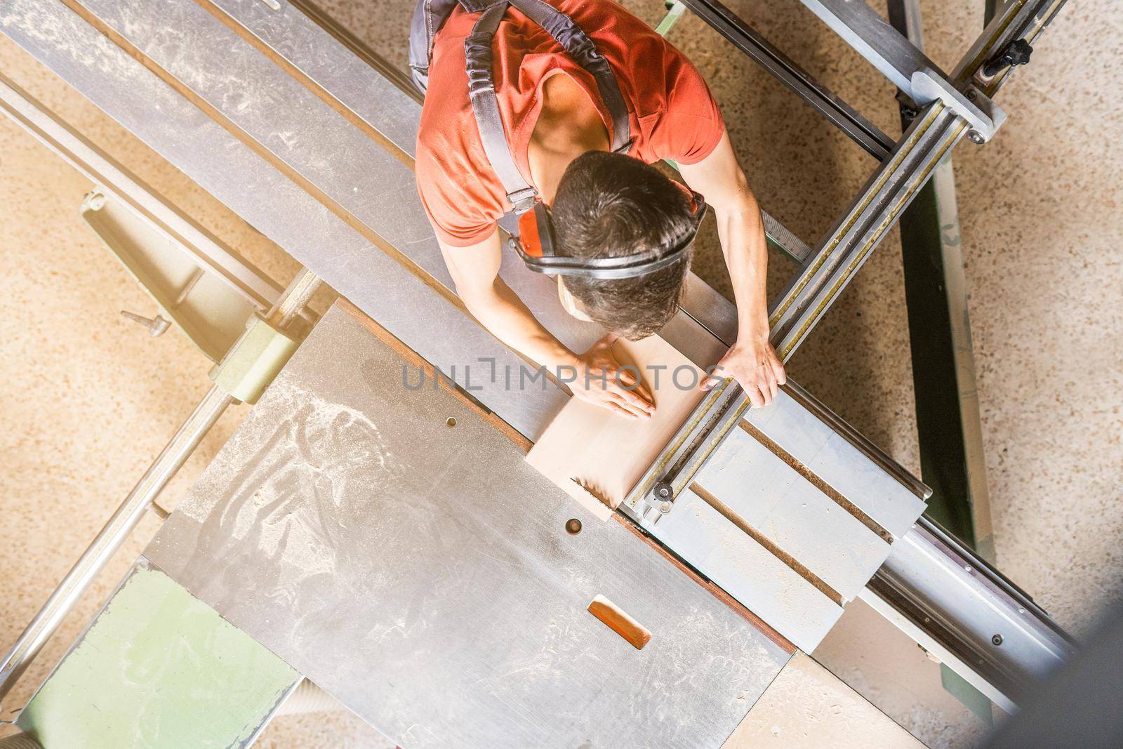 From above of anonymous male carpenter in protective headphones sawing wooden plank at squaring machine while working in professional joinery