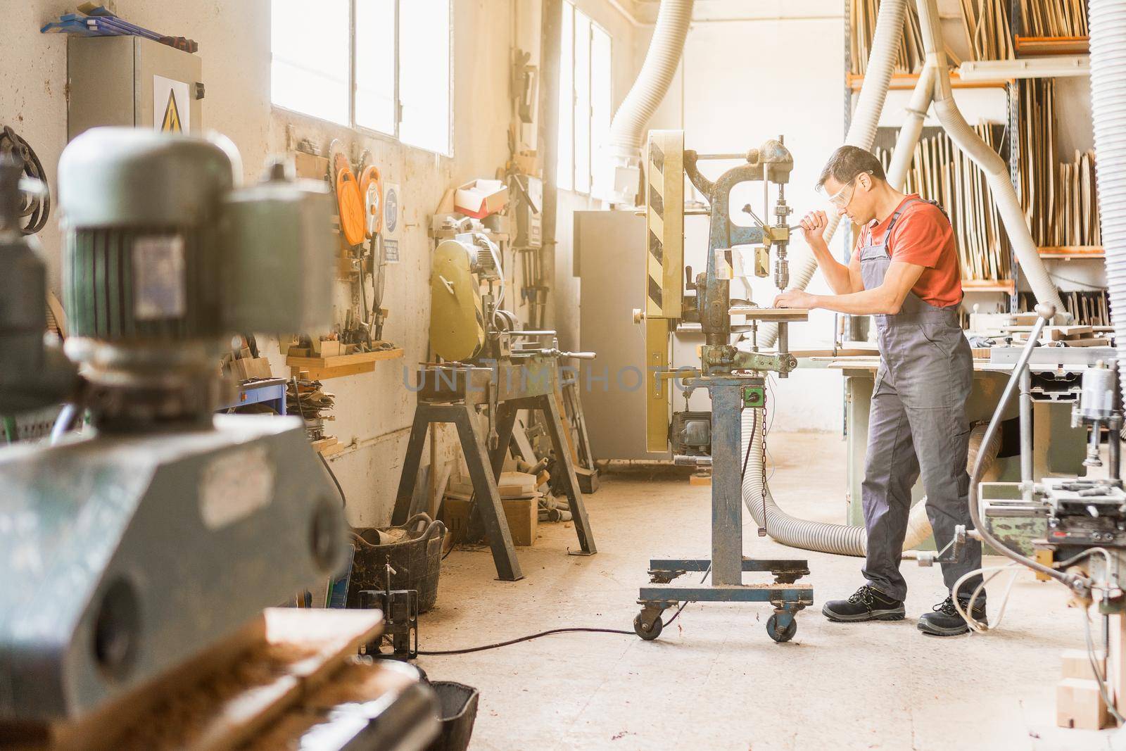 Full body side view of professional male carpenter cutting hole into wooden plank while working on drilling steel machine in joinery