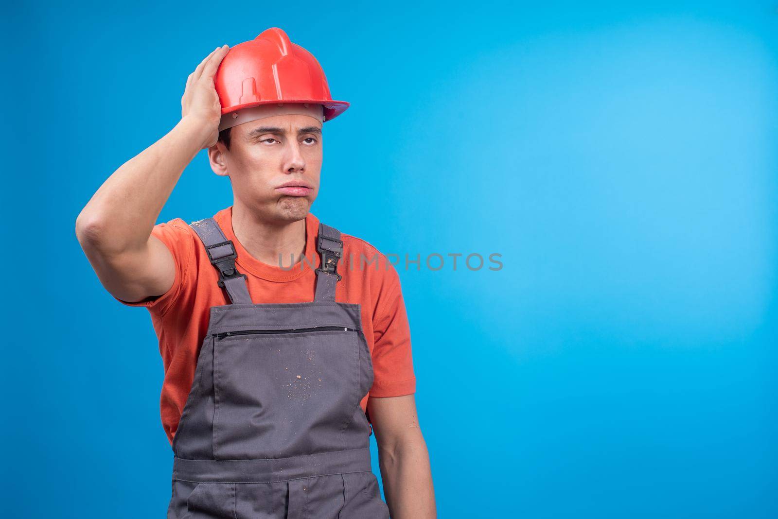 Exhausted man in builder workwear touching protective helmet and exhaling while taking break during work against blue background