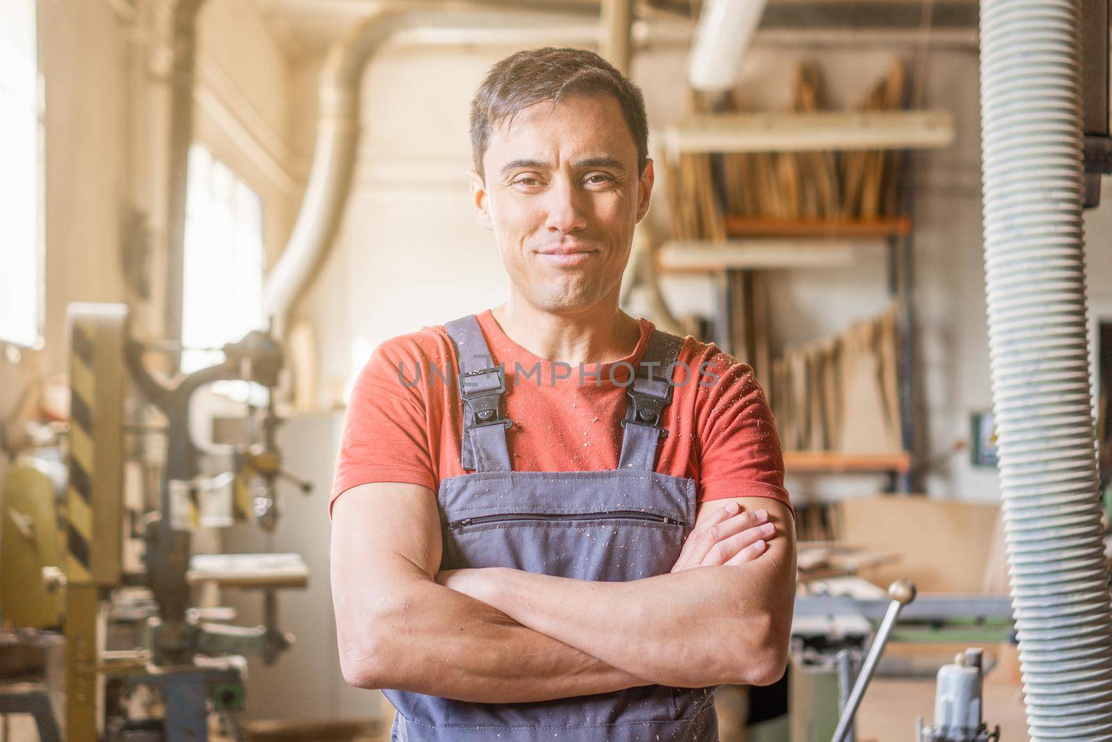 Content male woodworker in overall looking at camera with crossed arms while standing in professional light carpentry with various equipment