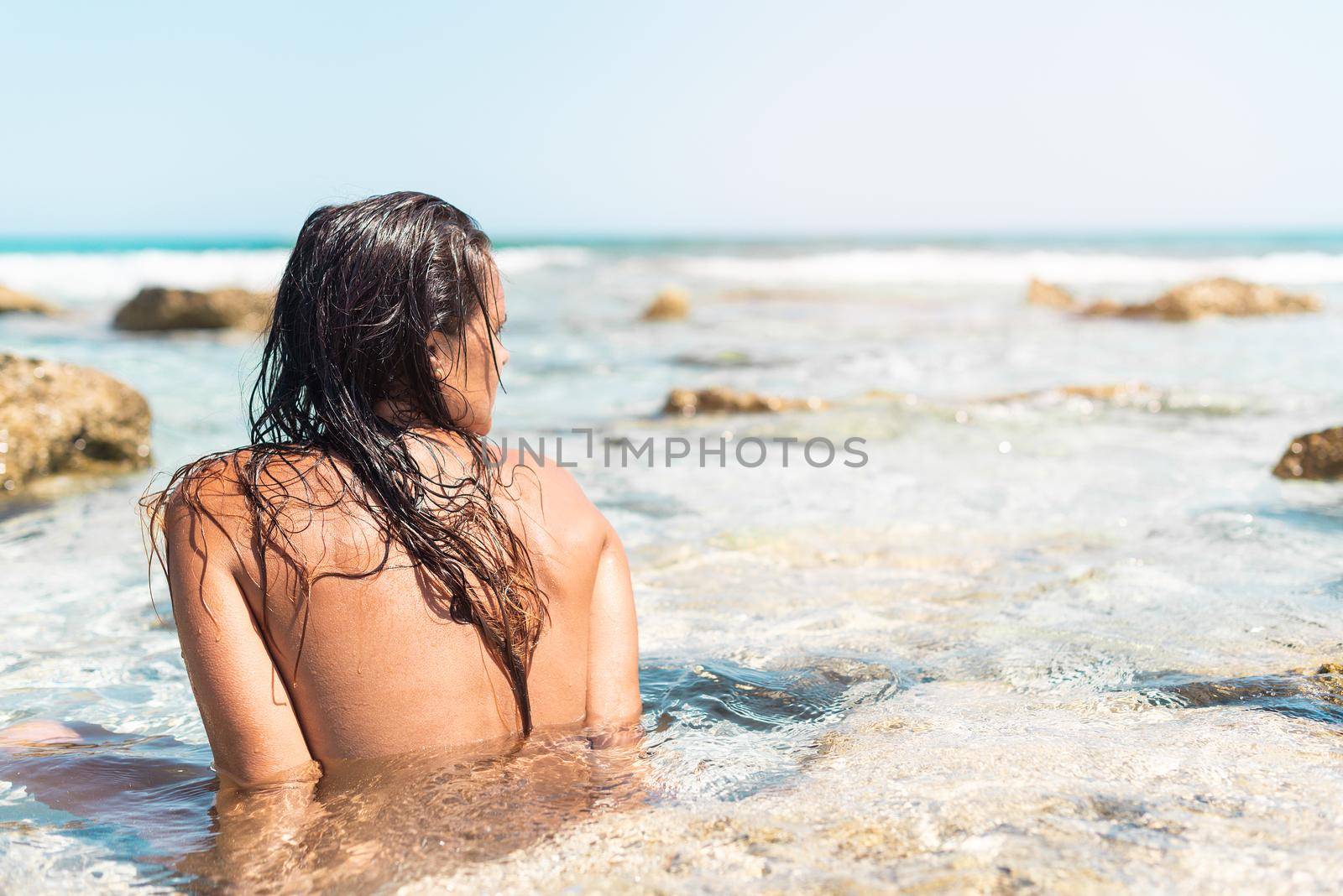 Back view of faceless nude female with wet hair sitting near rock in sea water and enjoying sunny day on Formentera island