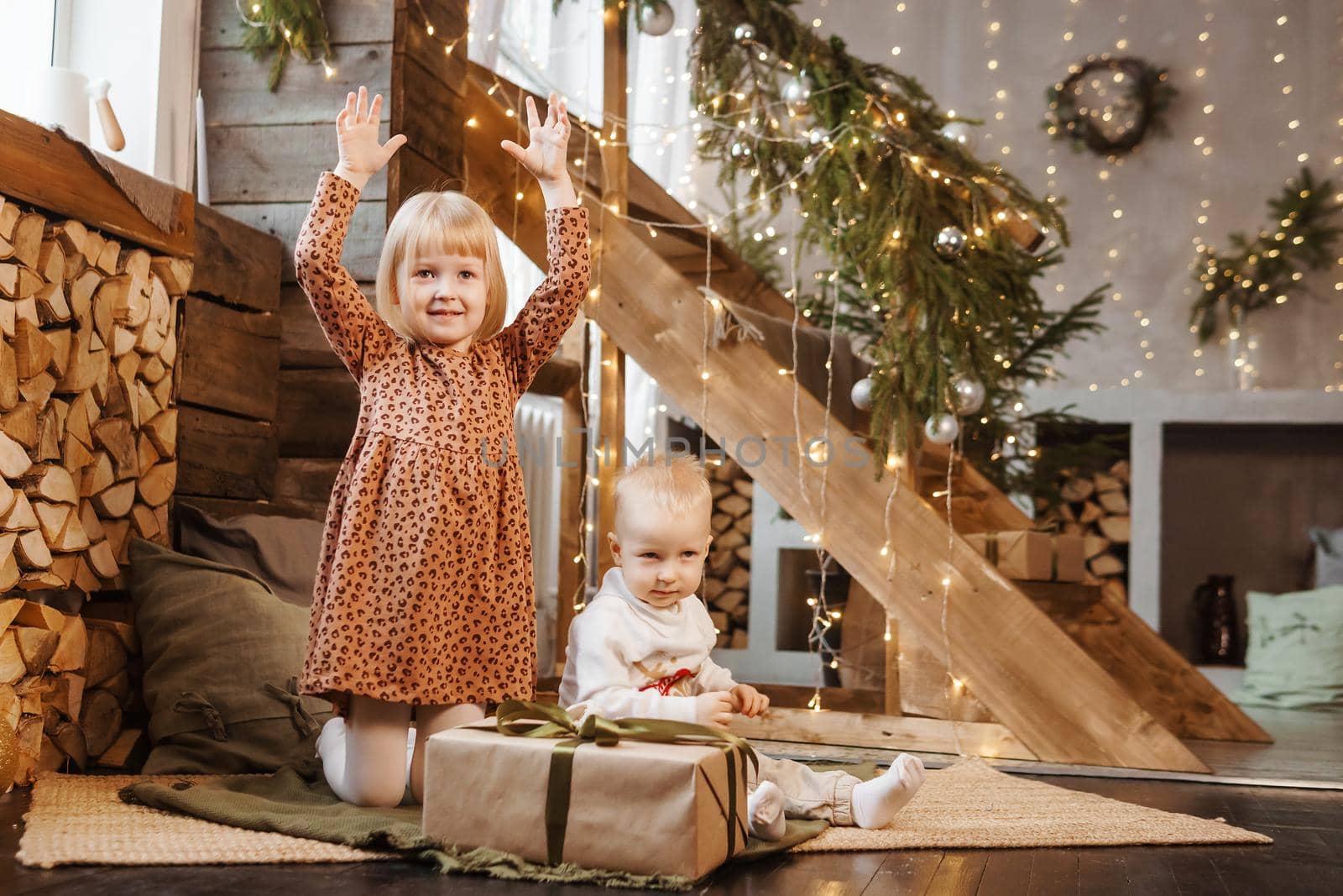 Little brother and sister play on Christmas eve in a beautiful house decorated for the New Year holidays. Children are playing with a Christmas gift. Scandinavian-style interior with live fir trees and a wooden staircase.