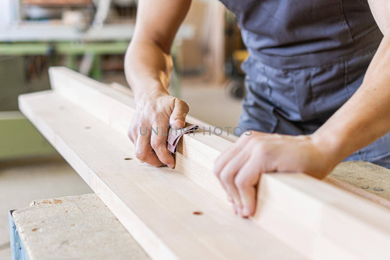 Faceless anonymous carpenter processing long wooden desk with sandpaper, while working in joinery on blurred background