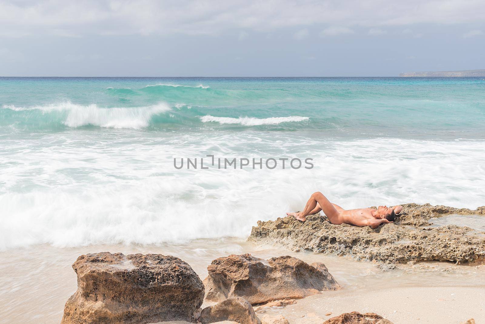 Nude man with hands behind head lying on rocks and sunbathing while resting on beach near stormy sea on Formentera island in Spain