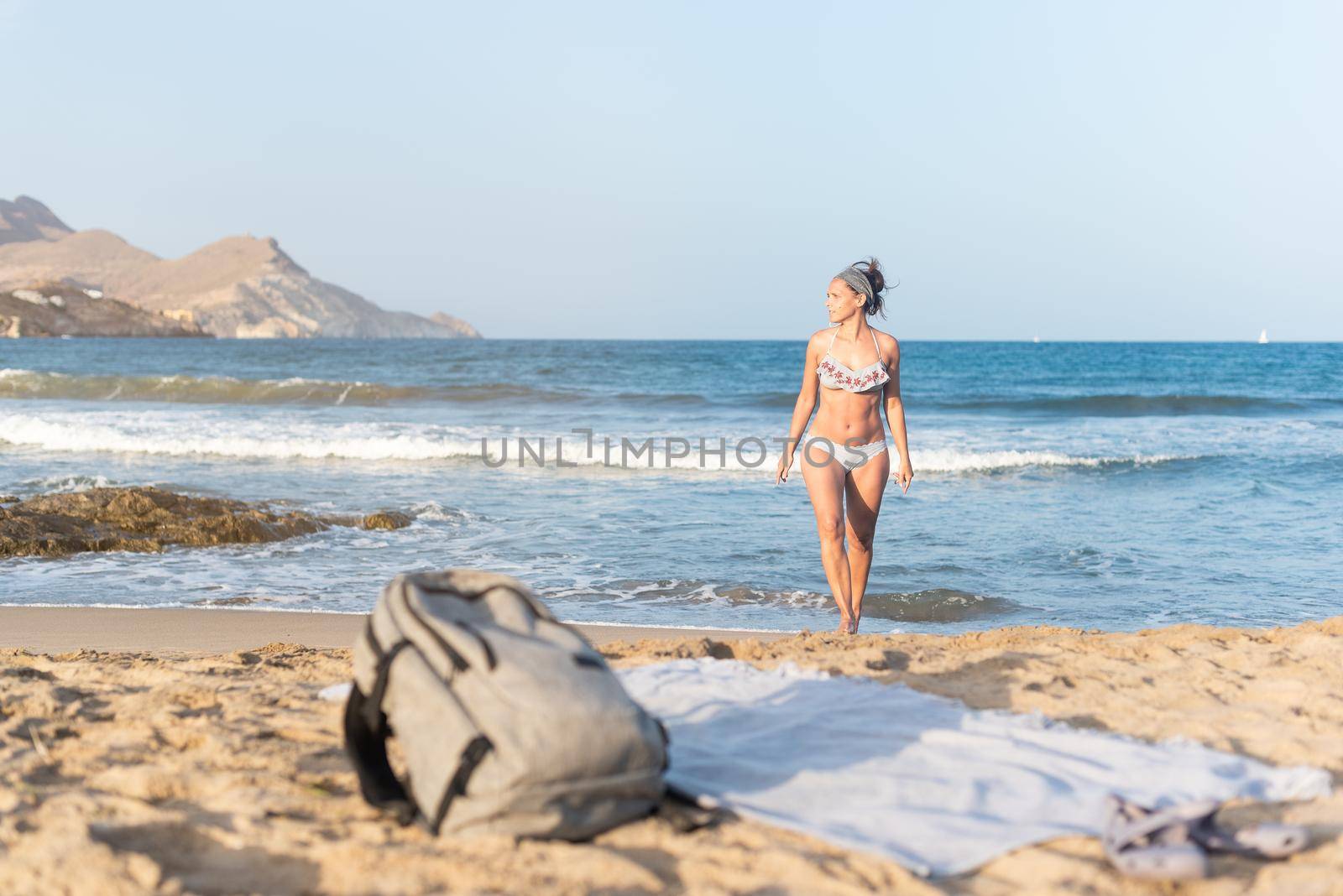 Full length of slim female tourist in swimwear walking near waving ocean towards backpack and towel on sandy beach, and looking away during vacation in Almeria Andalusia Spain