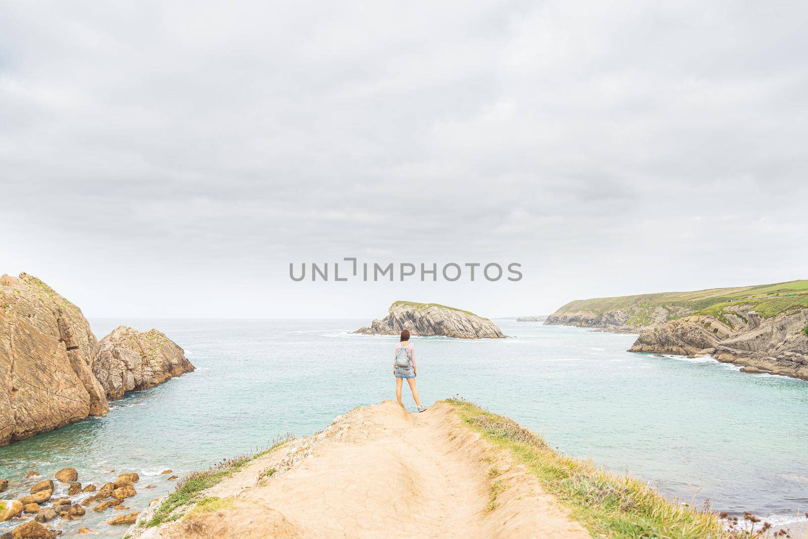 Back view of unrecognizable woman with backpack admiring view of blue sea, while standing on sandy coast near rocky formation on Arnia beach in Cantabria in Spain in