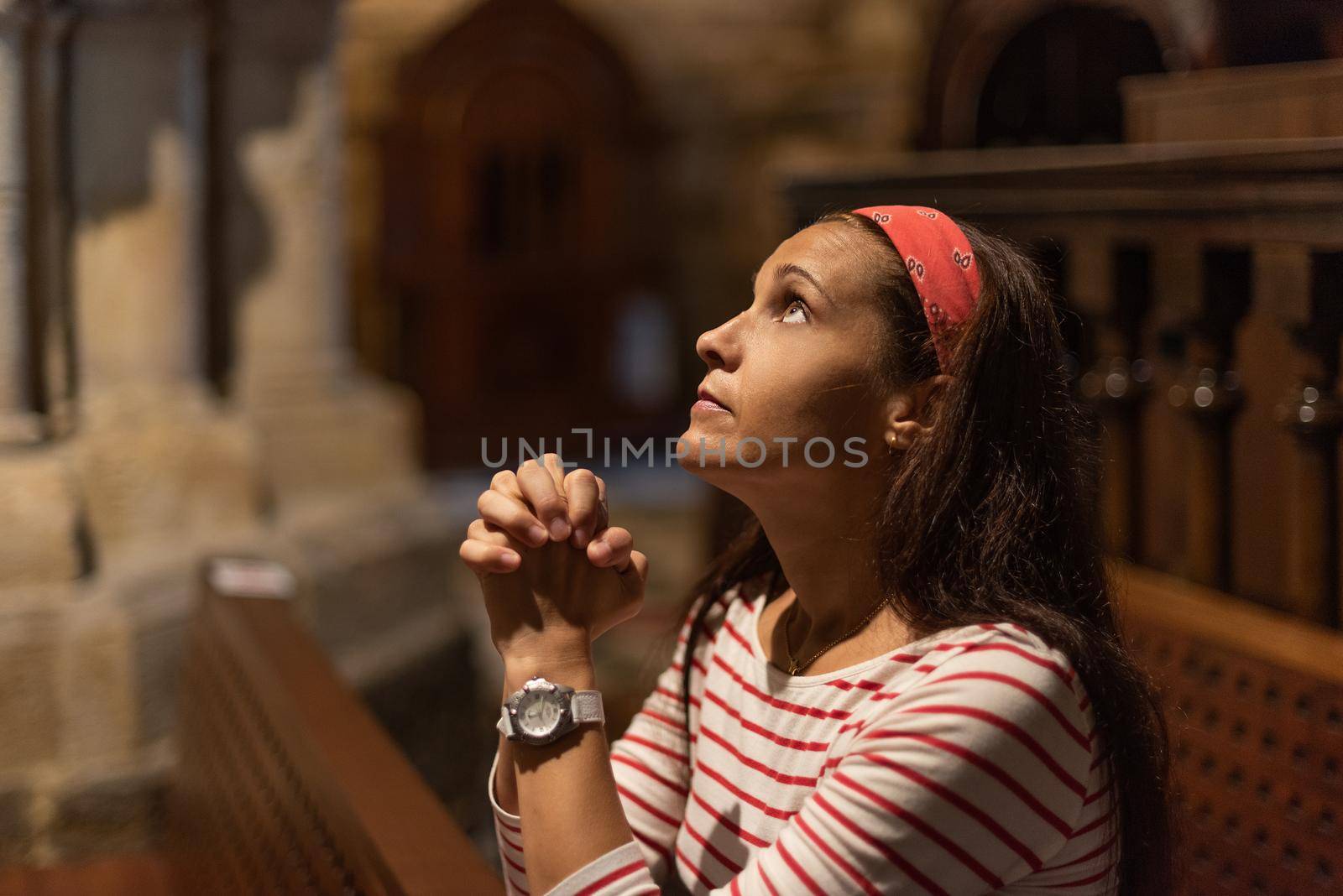 Side view of serious Hispanic female with long dark hair giving prayer with clasped hands, while sitting on bench in old Catholic church and looking up