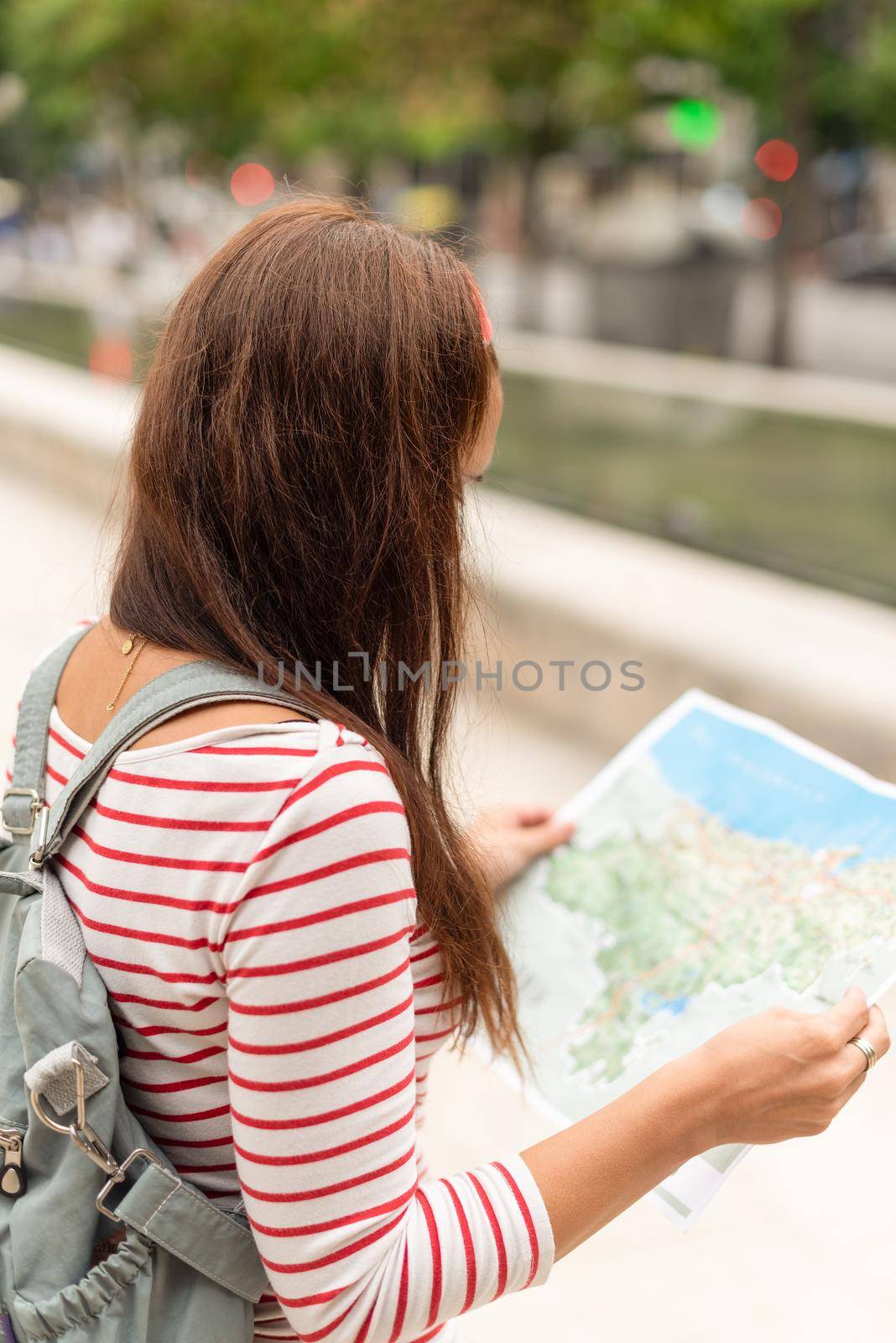 Back view of unrecognizable female tourist with long dark hair and backpack exploring map while walking in city in Spain in summer day