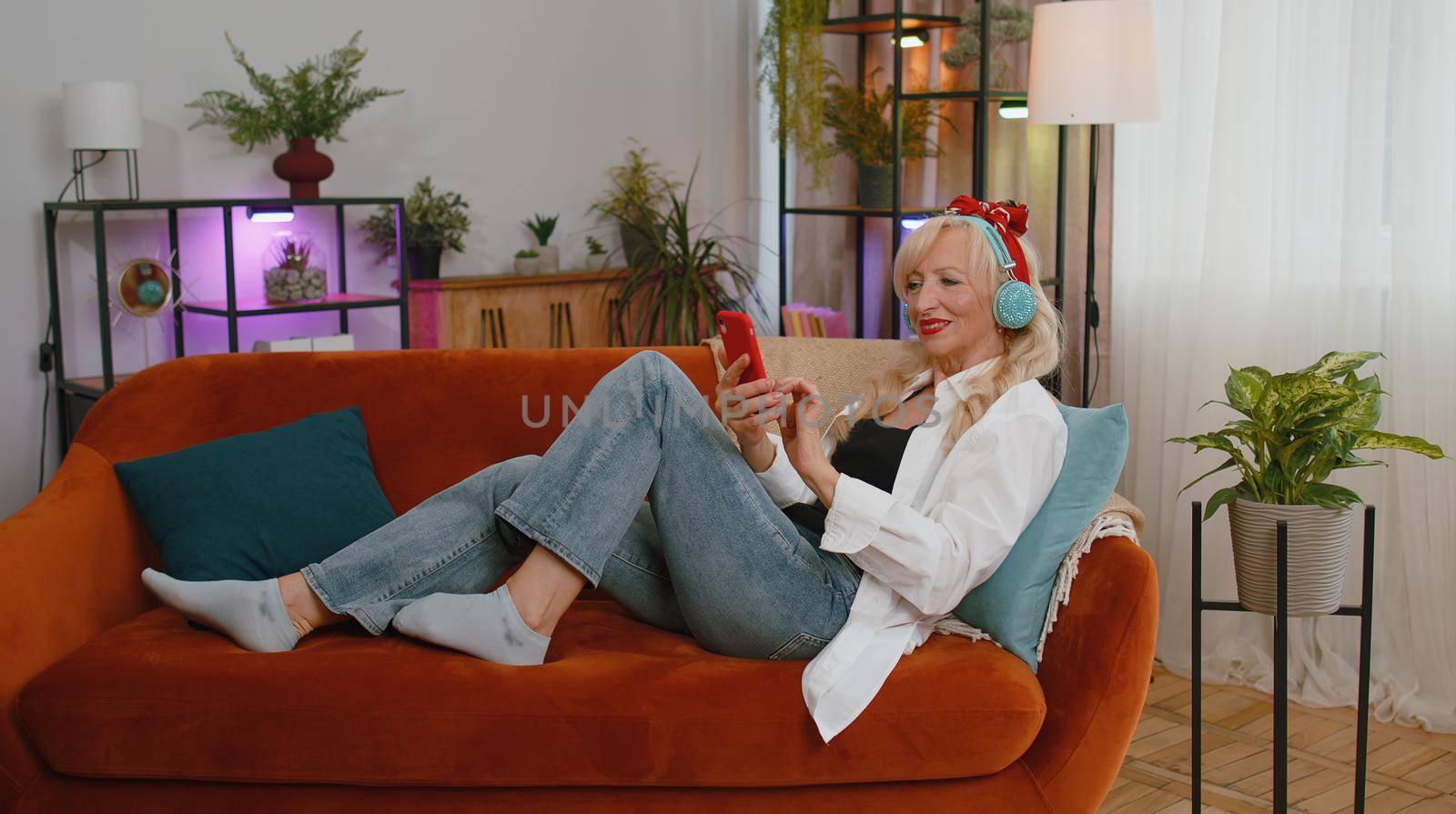 Happy relaxed overjoyed senior woman in wireless headphones dancing on couch at home choosing listening favorite energetic disco rock n roll music in smartphone entertaining. People weekend activities