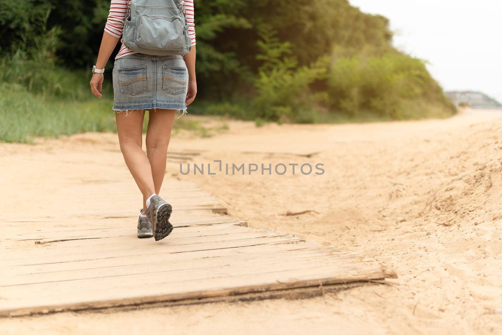 Back view of crop unrecognizable female tourist in denim skirt and backpack strolling along wooden boardwalk.