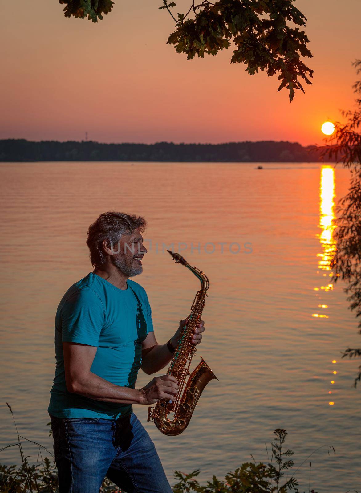 An adult man plays the saxophone at sunset by the river in the evening.