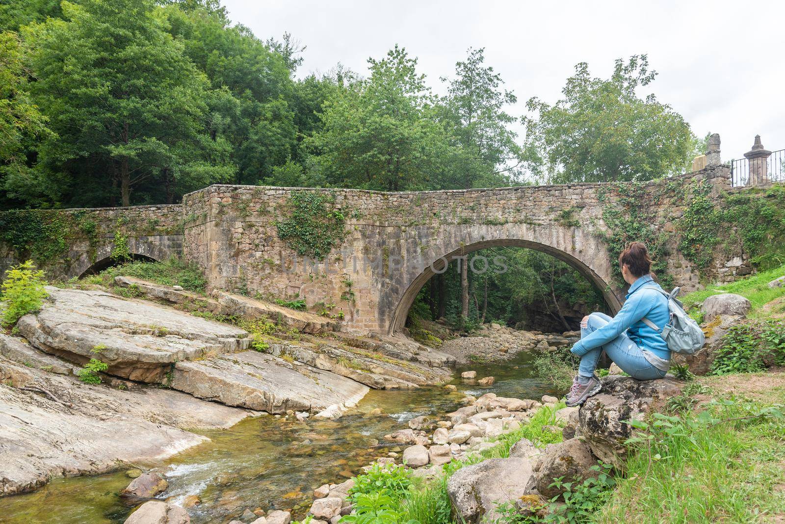 Side view full body of unrecognizable woman sitting on stones and admiring view of old arched stone bridge over narrow creek, in Barcena Mayor in Cantabria in Spain