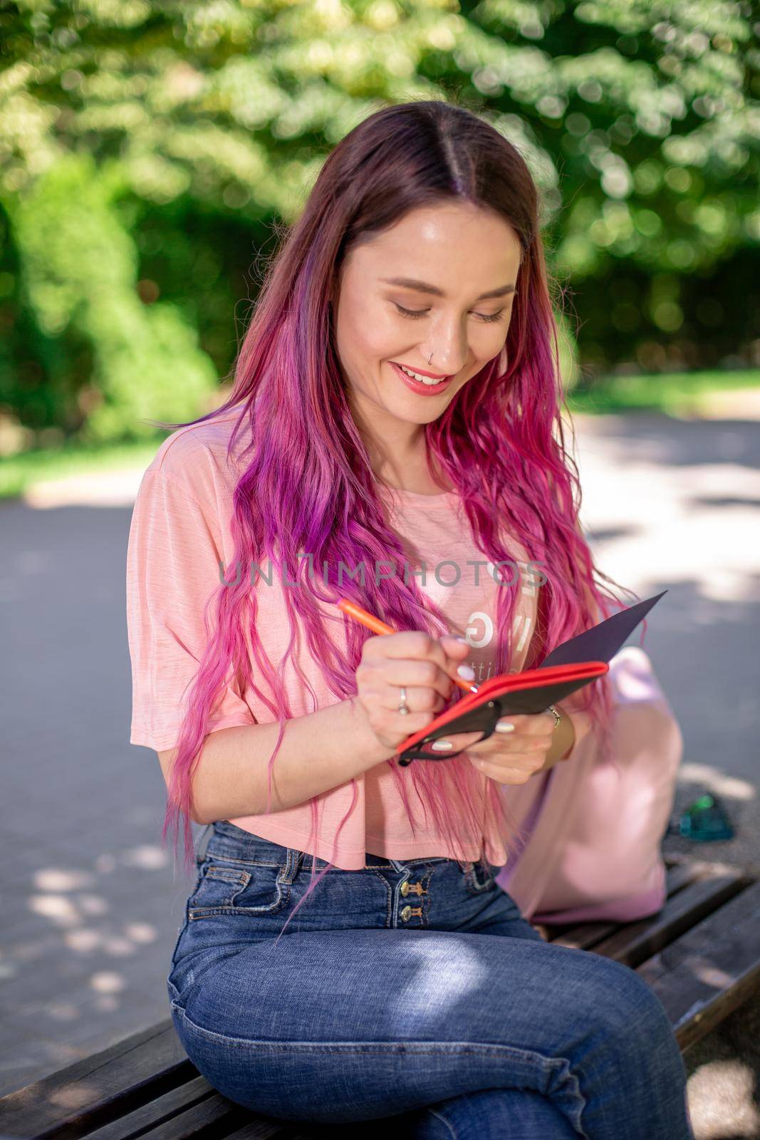 Woman writing in a notebook sitting on a wooden bench in the park. Girl working outdoors on portable computer, copy space. Technology, communication, freelance and remote working concept.