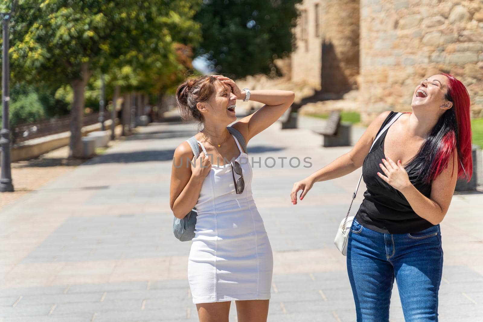 Delighted female friends in summer clothes walking on street in Avila city and laughing while having fun together on sunny day
