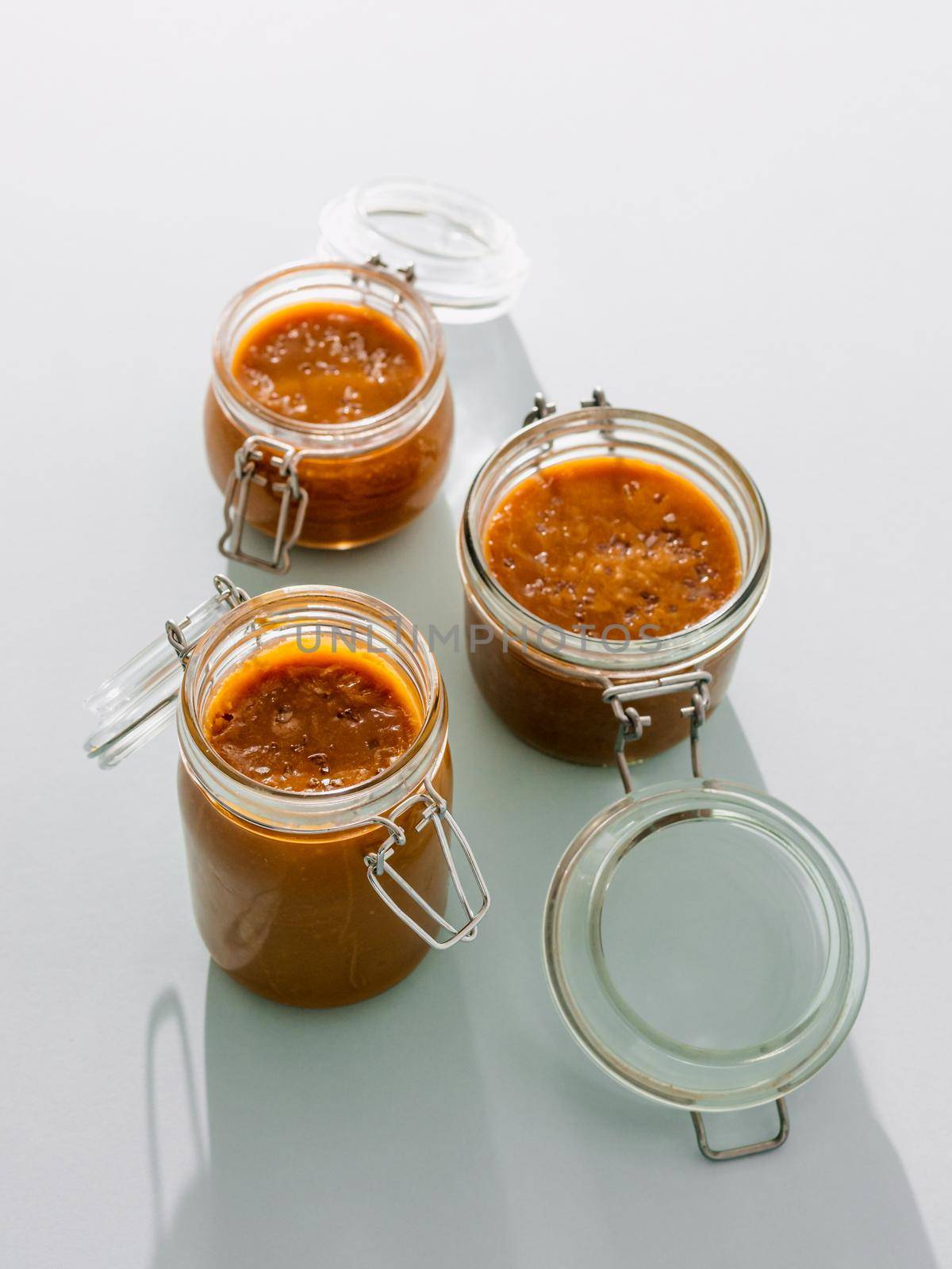 Salted caramel in glass jars. Brown caramel or condensed milk with sea salt crystalls, shoot in bright or hard light. Light neutral background. Vertical