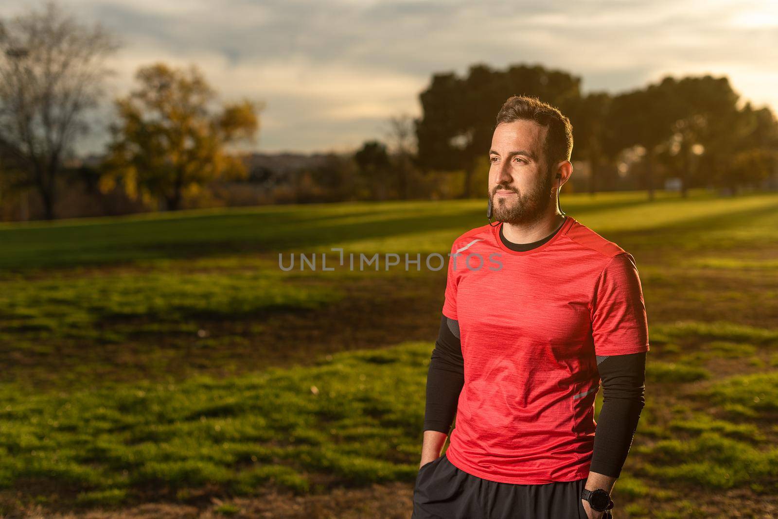 Bearded male runner standing with hands in pockets of shorts and listening to music during break in jogging training