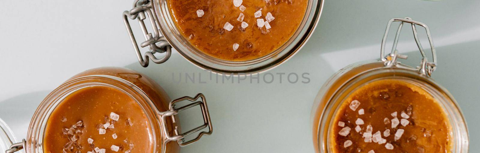 Salted caramel in glass jars, banner horizontal, top view. Brown caramel or condensed milk with sea salt crystalls, shoot in bright or hard light. Light neutral background