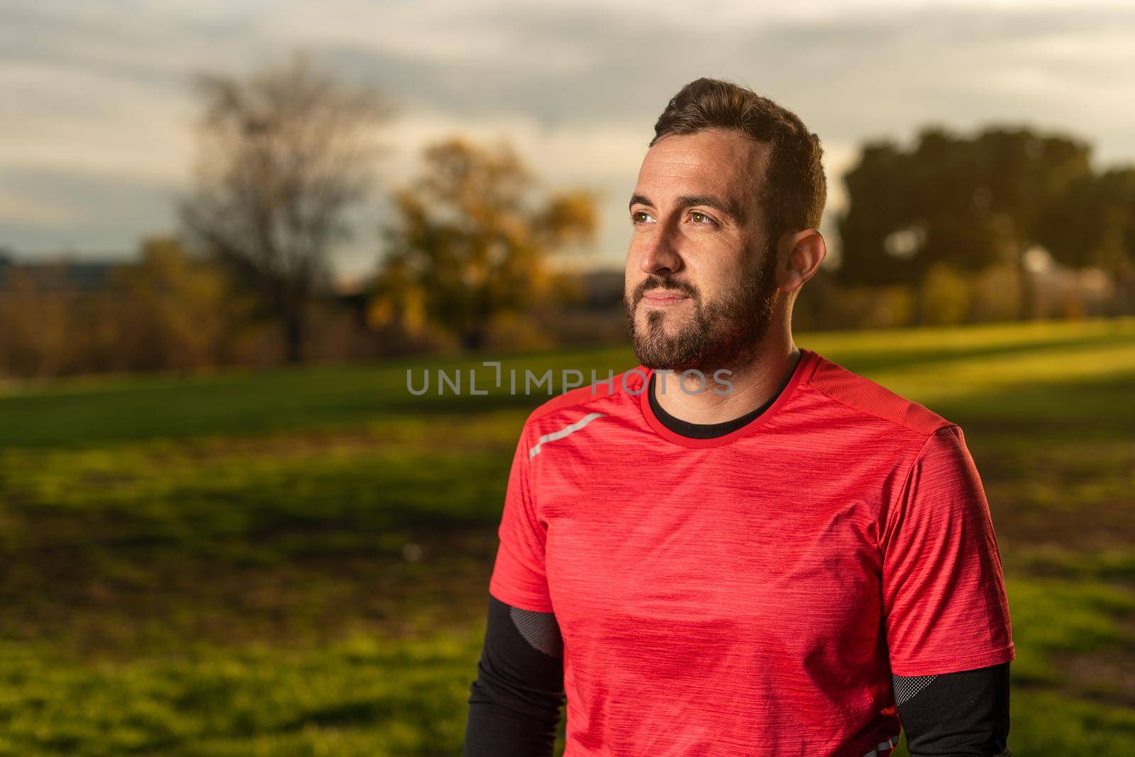 Male athlete in sportswear standing on grassy lawn during fitness training in park in evening and looking away with gentle smile