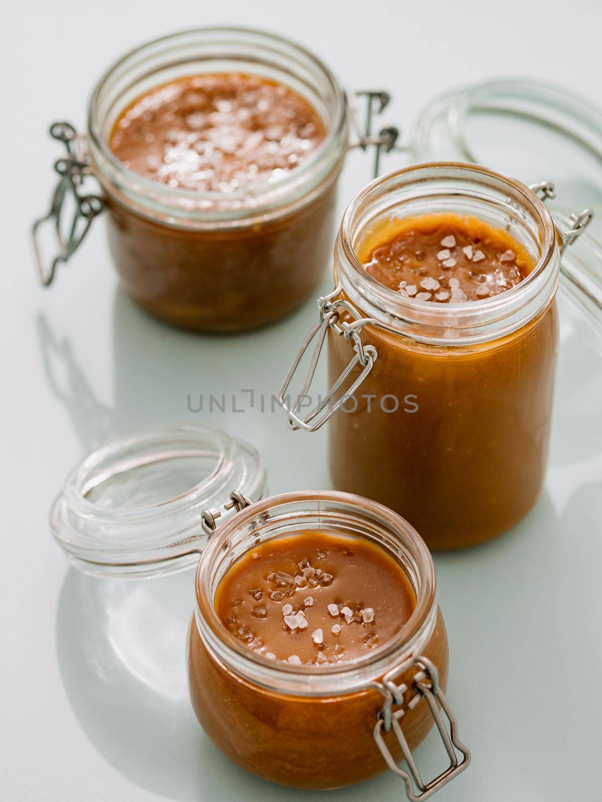 Salted caramel in glass jars. Brown caramel or condensed milk with sea salt crystalls, shoot in bright or hard light. Light neutral background. Vertical