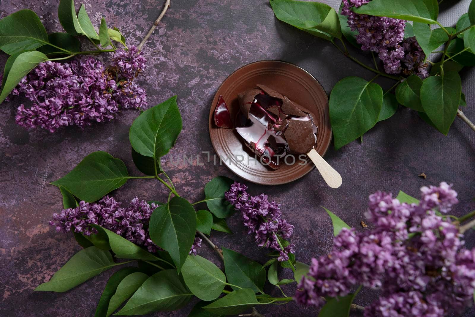 melted raspberry ice cream in chocolate on a wooden stick on a dark burgundy background with lilac flowers, a sweet dessert on a hot spring day, flat lay food. High quality photo