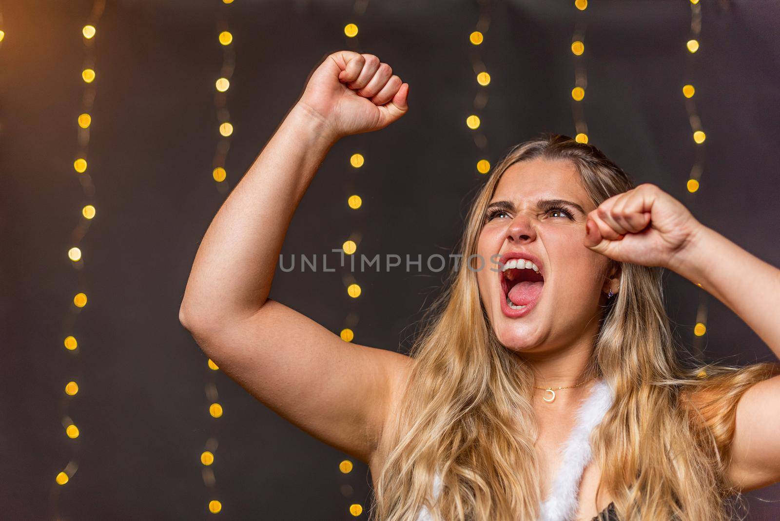 A euphoric Caucasian woman screaming and raising her hands with clenched fists against a background of blurred lights.