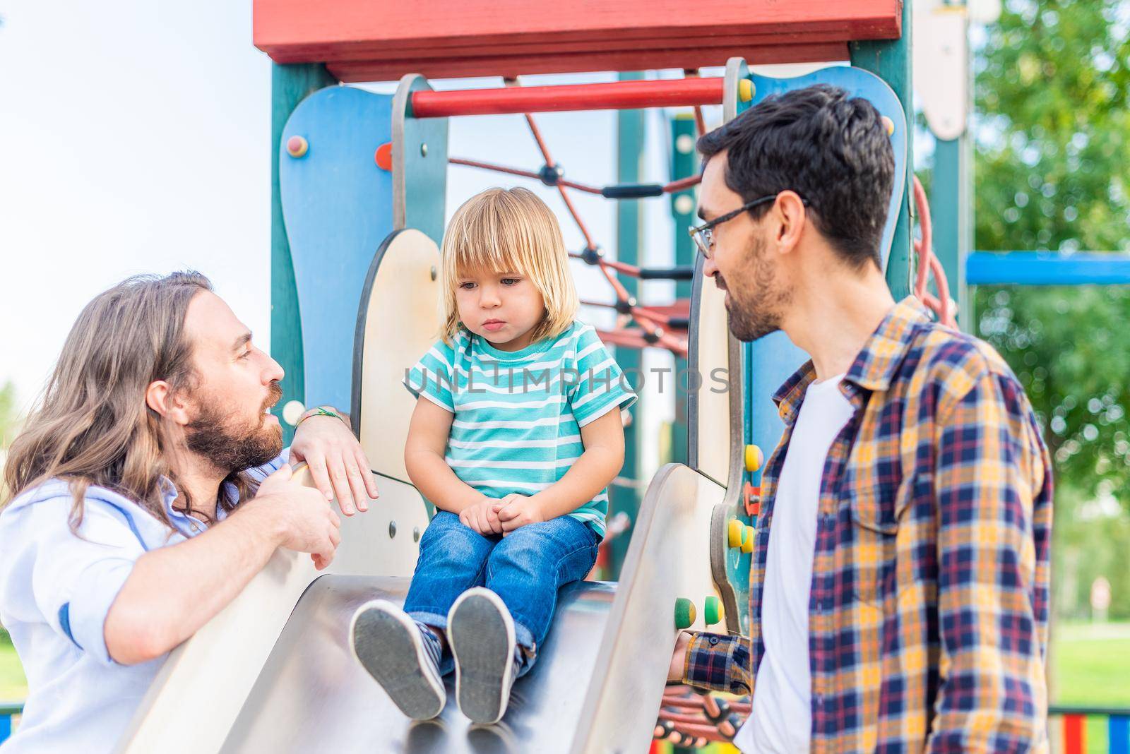 little blond boy on top of a slide looking down with a fear of heights cared for by his two adoptive parents.