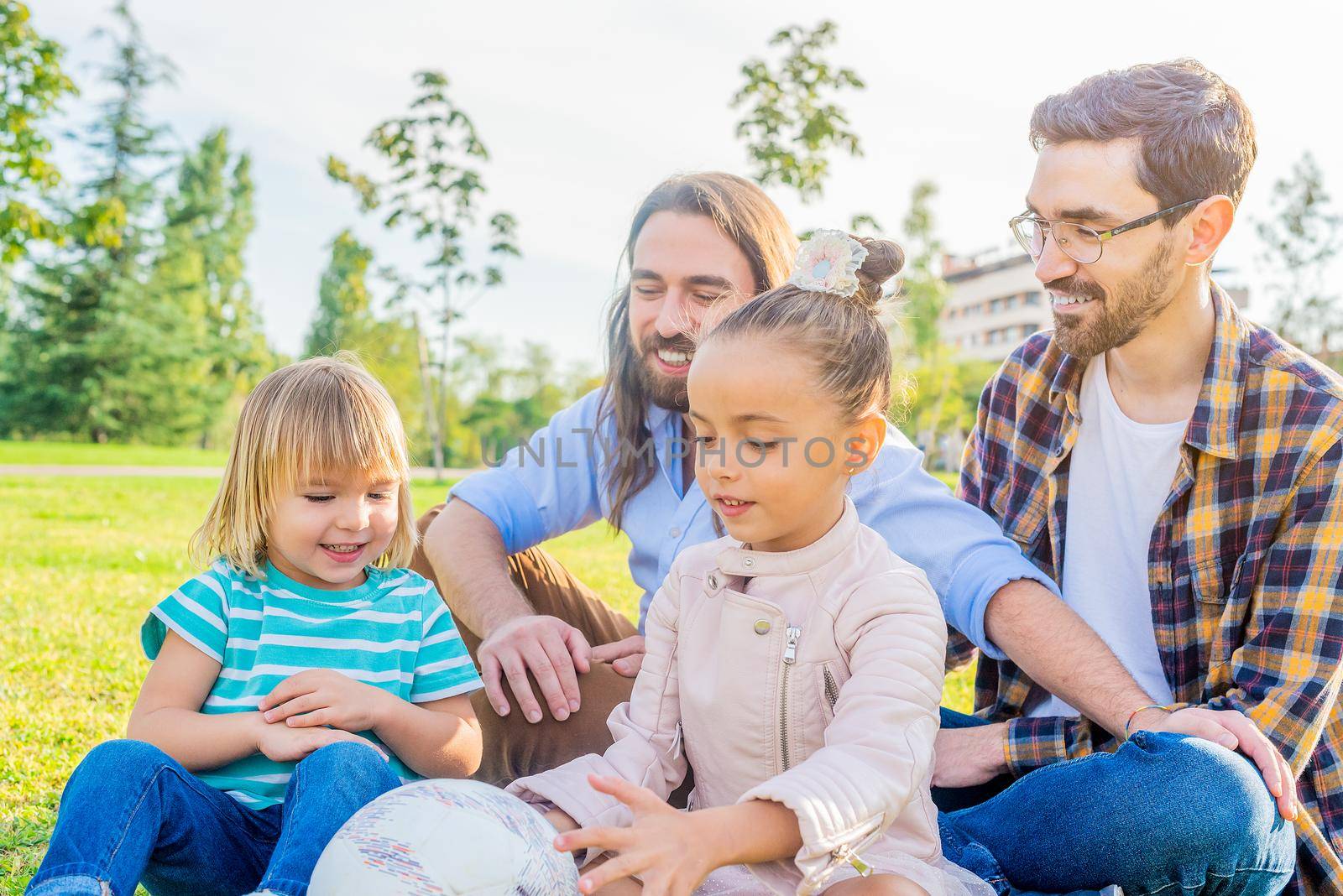 Portrait of a gay male couple with their two children sitting on the grass in a park having a good time as a family.