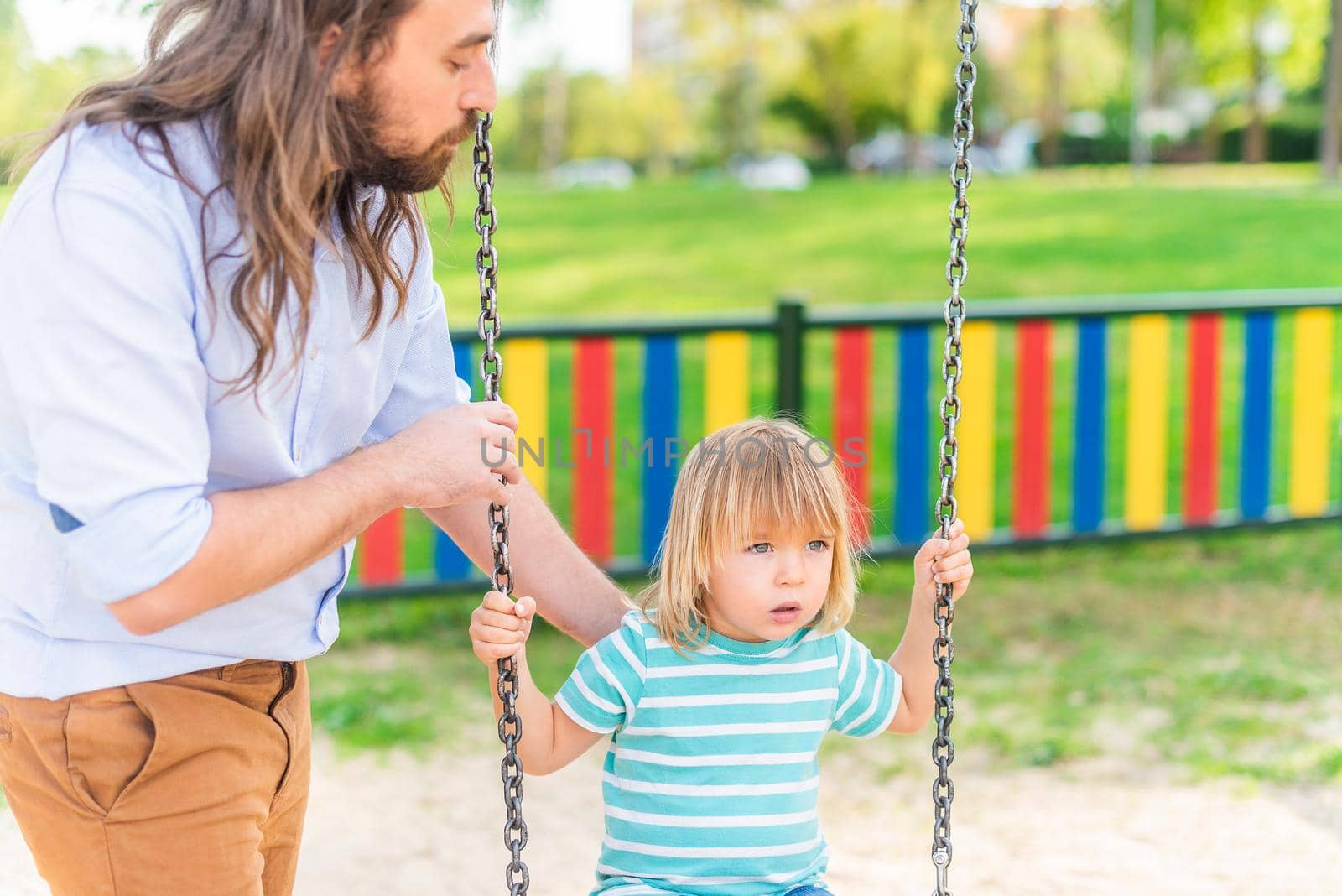 A young man playing with his adopted son on the swing in a playground in a sunny day.