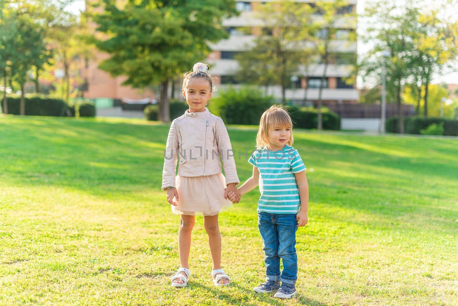 Front view of a little girl looking at the camera holding hands with her little brother standing in the park.