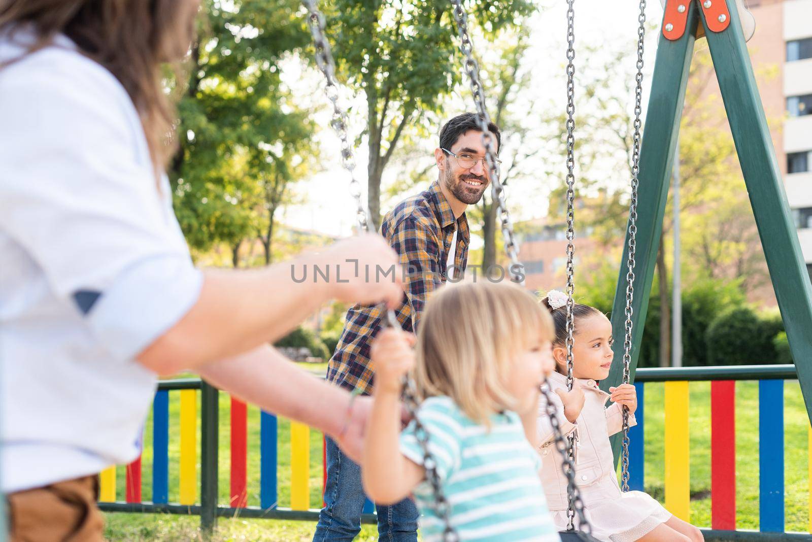 Side view of a happy young gay couple looking at each other playing with their children on the swing.