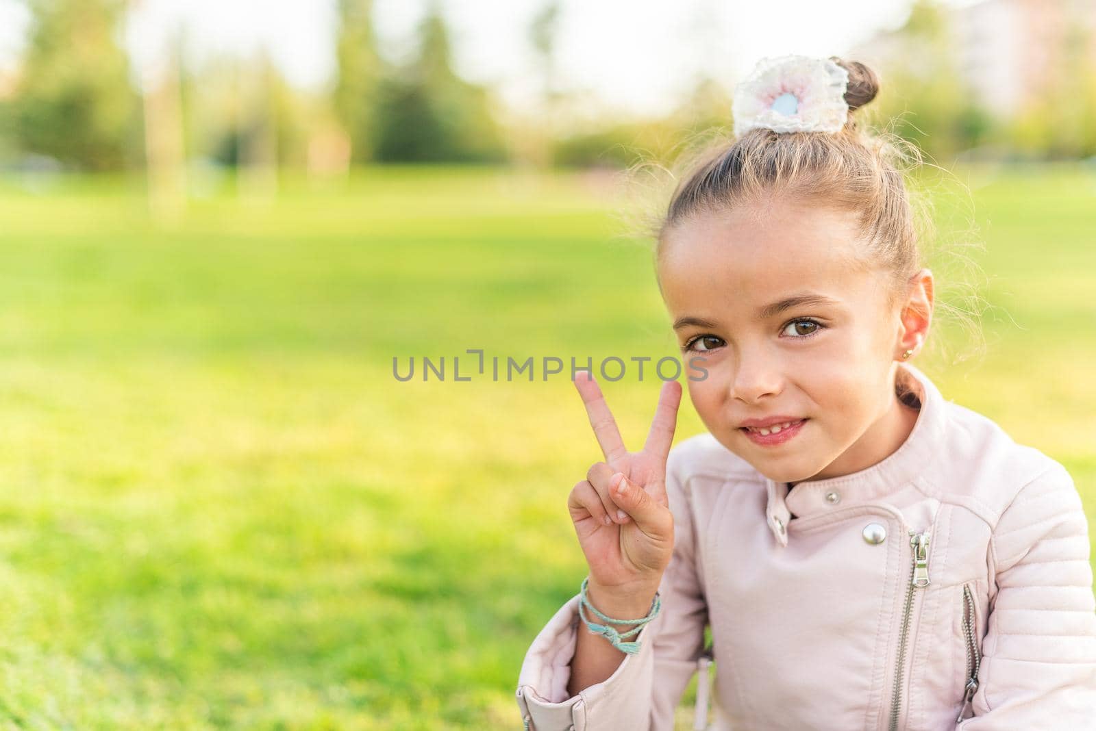 Portrait of a little girl sitting on grass in a park looking at camera and showing peace or victory sign with copy space