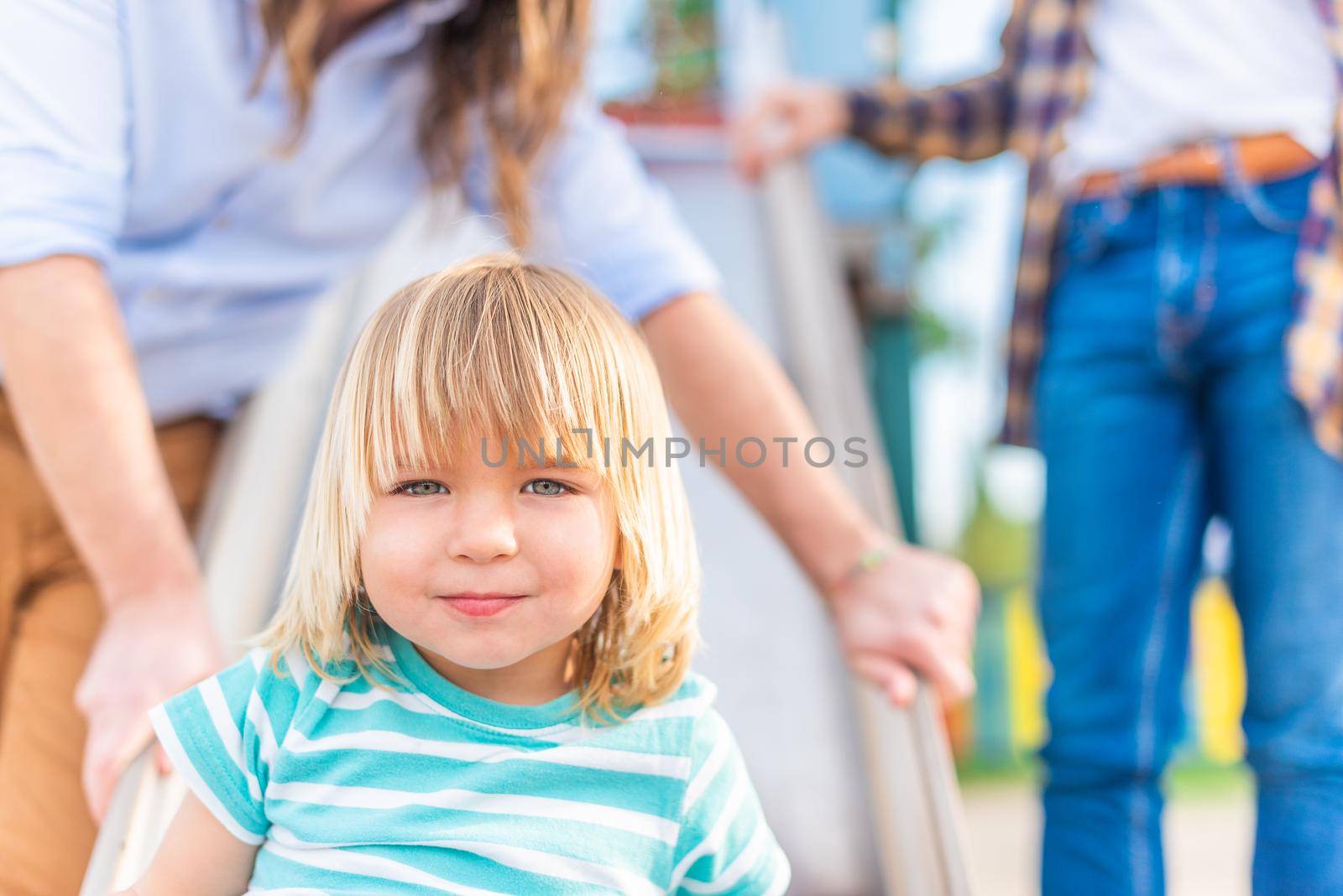 Portrait of a happy blond boy looking at the camera sitting on a playground slide with his two unrecognizable dads behind him.