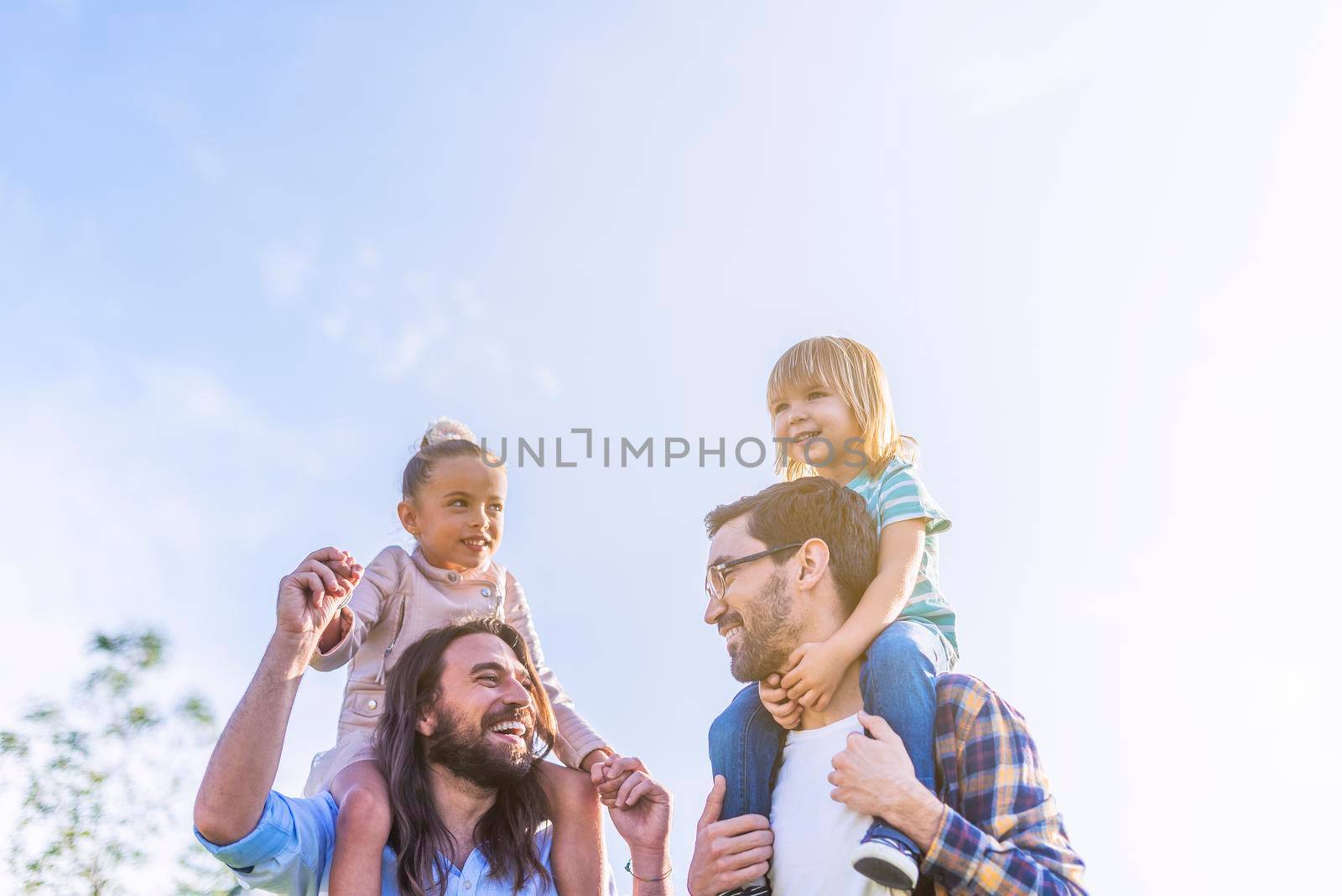 Front view of a young gay male couple holding their young children on their shoulders in the park with copy space