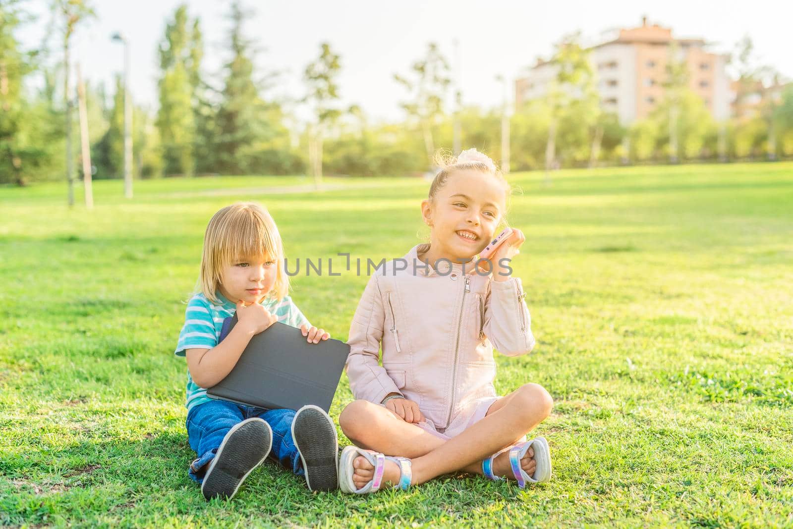 Front view of a little girl talking on a cell phone sitting on the grass with her little brother holding a digital tablet