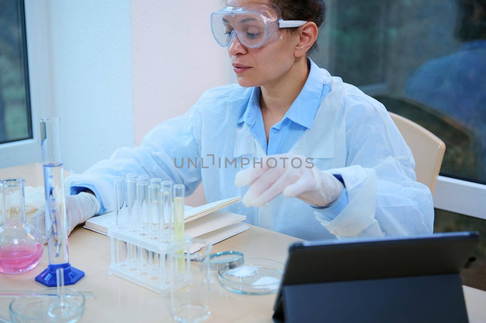 Charming Latin American woman, a healthcare and laboratory worker, pharmacist, pharmacologist, experienced scientist wearing lab coat and safety googles, conducting scientific chemical experiment