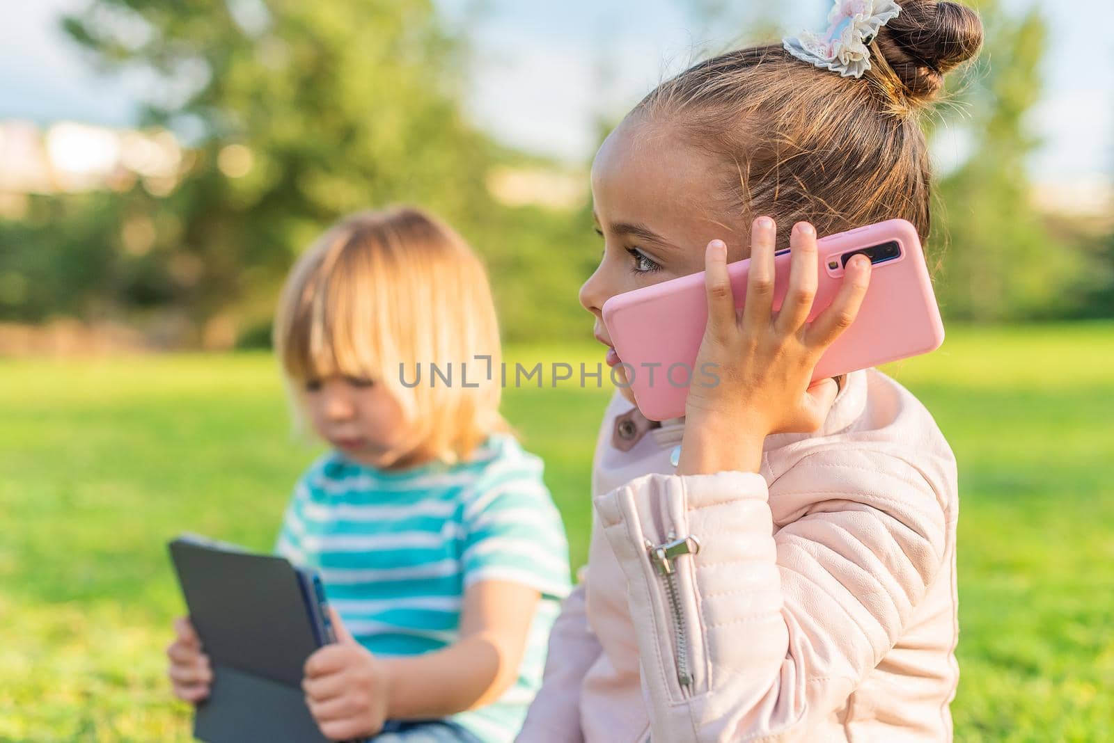 Side view of a little girl talking on a smart phone sitting on the grass with her little brother holding a digital tablet