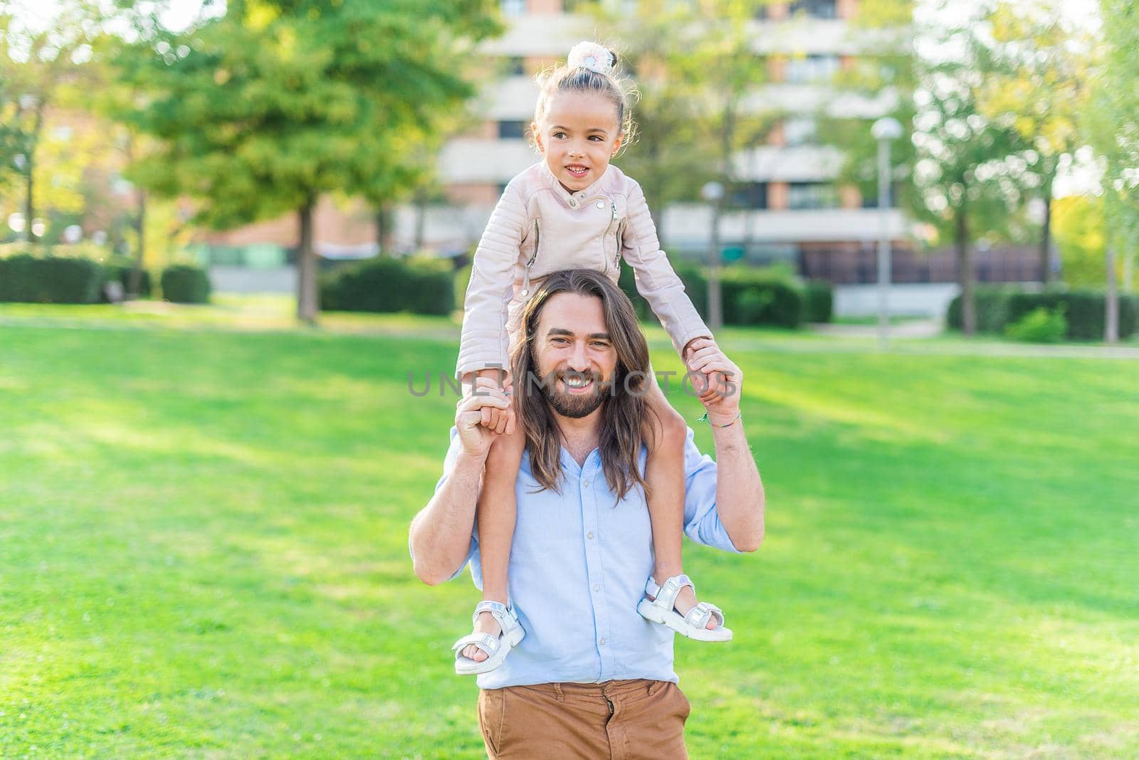 Front view of a happy young father holding her little daughter on his shoulder in park