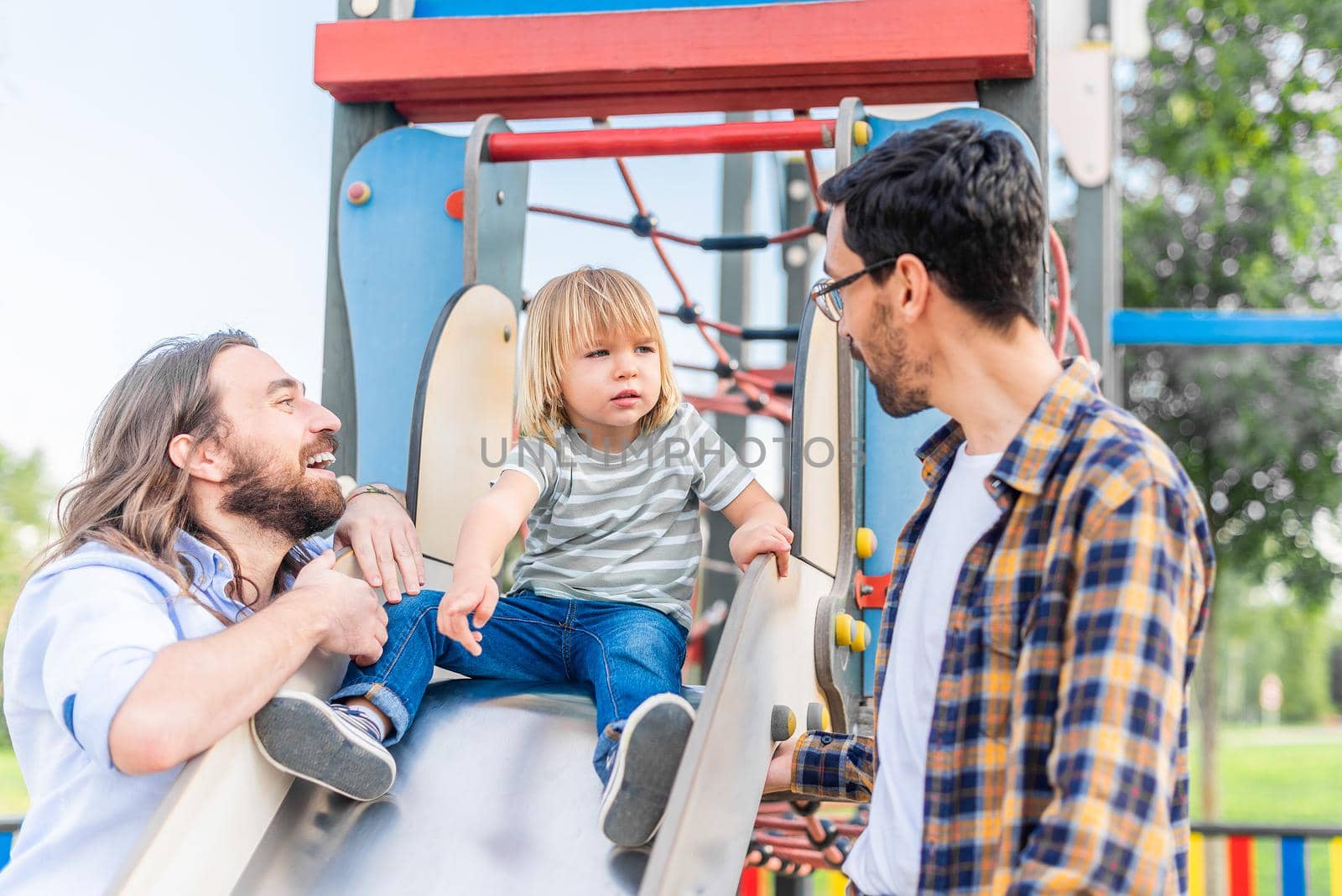 Front view of a little blond boy on top of a slide playing on it for the first time with his two dads.