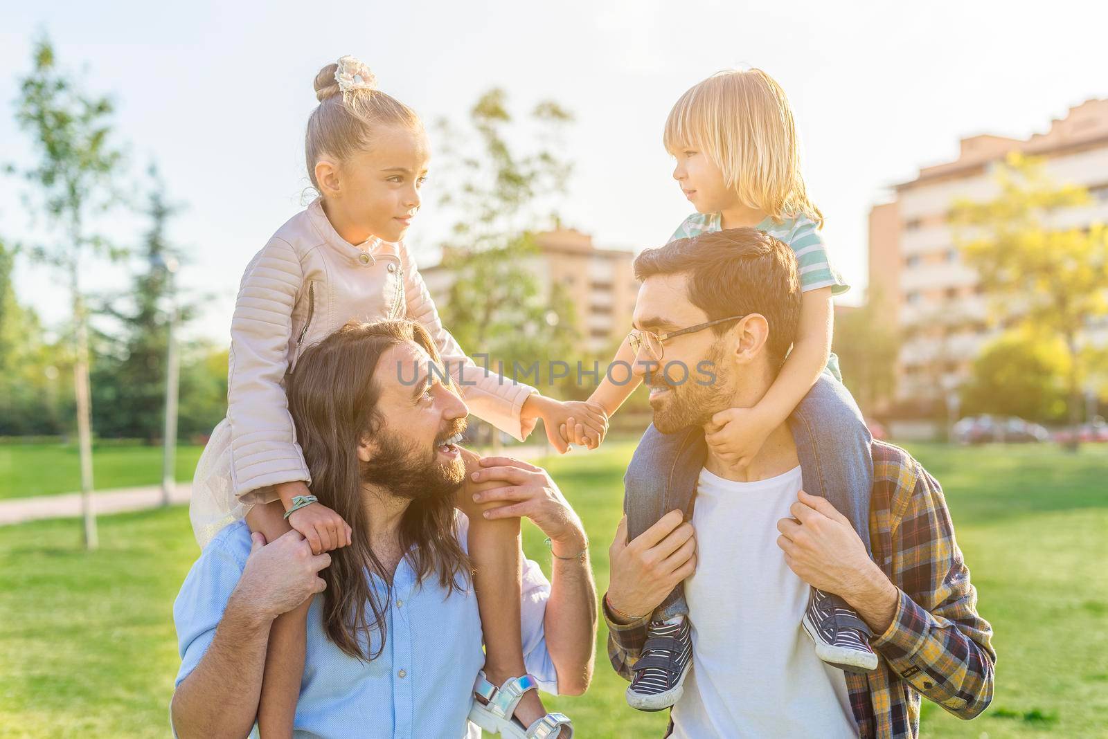 Front view of a young gay male couple holding on their shoulders their children holding hands in the park