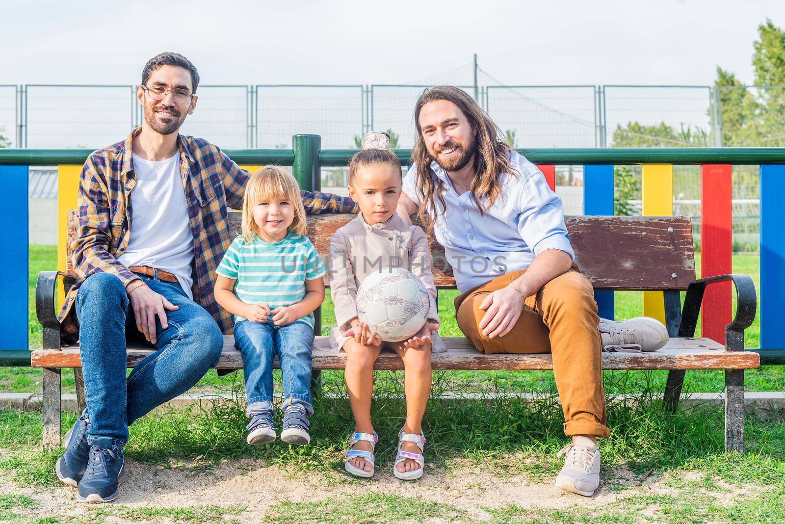 Portrait of gay male couple with their two children looking at camera sitting on a bench in the playground.