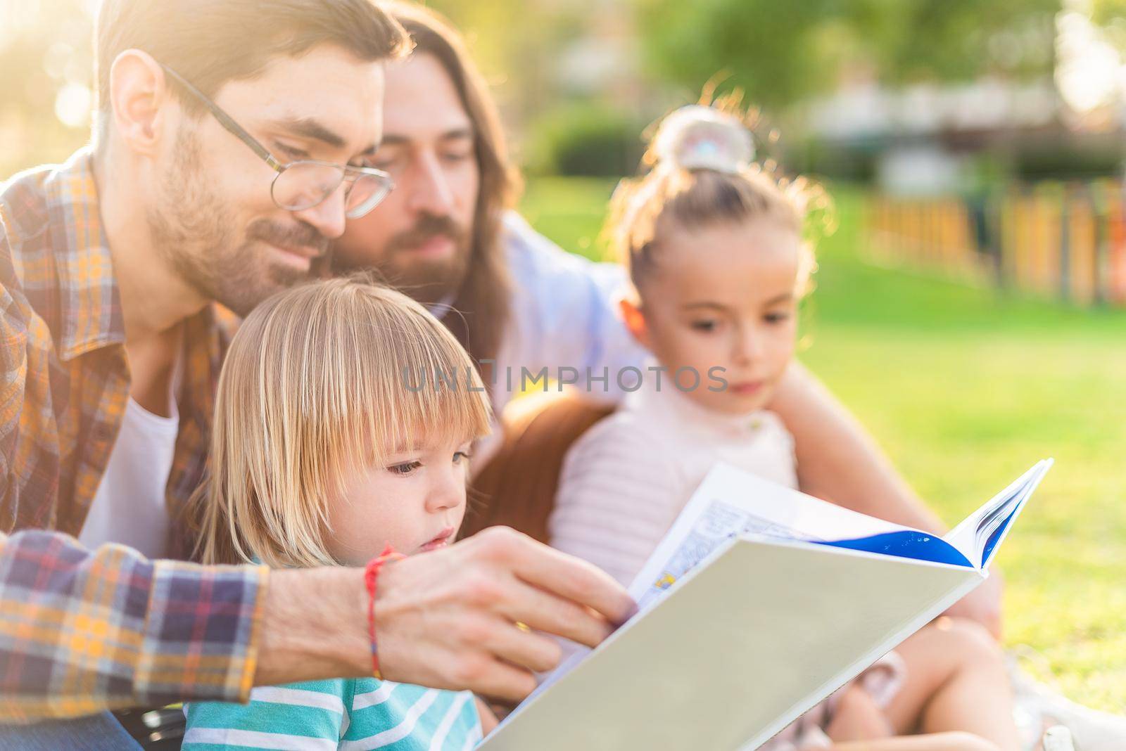 Side view of a young gay male couple with their adopted children in the park reading a children's book in a sunny day.