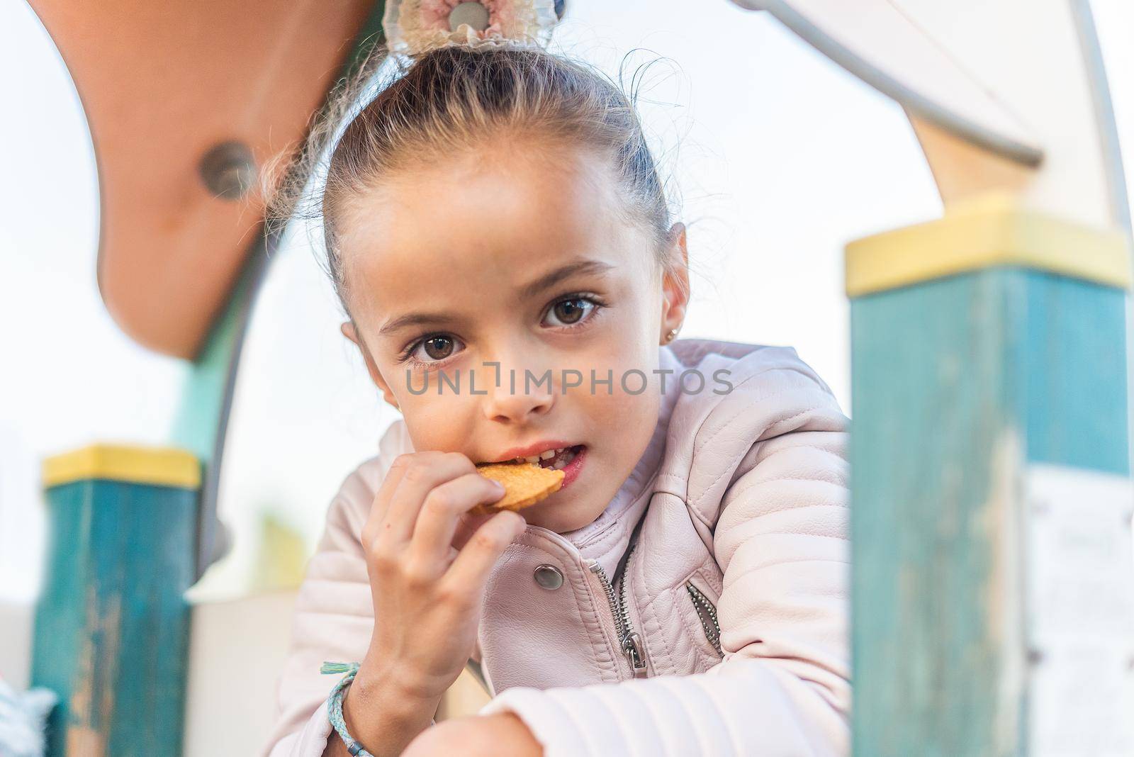 girl looking at camera eating a cookie in a playground by ivanmoreno