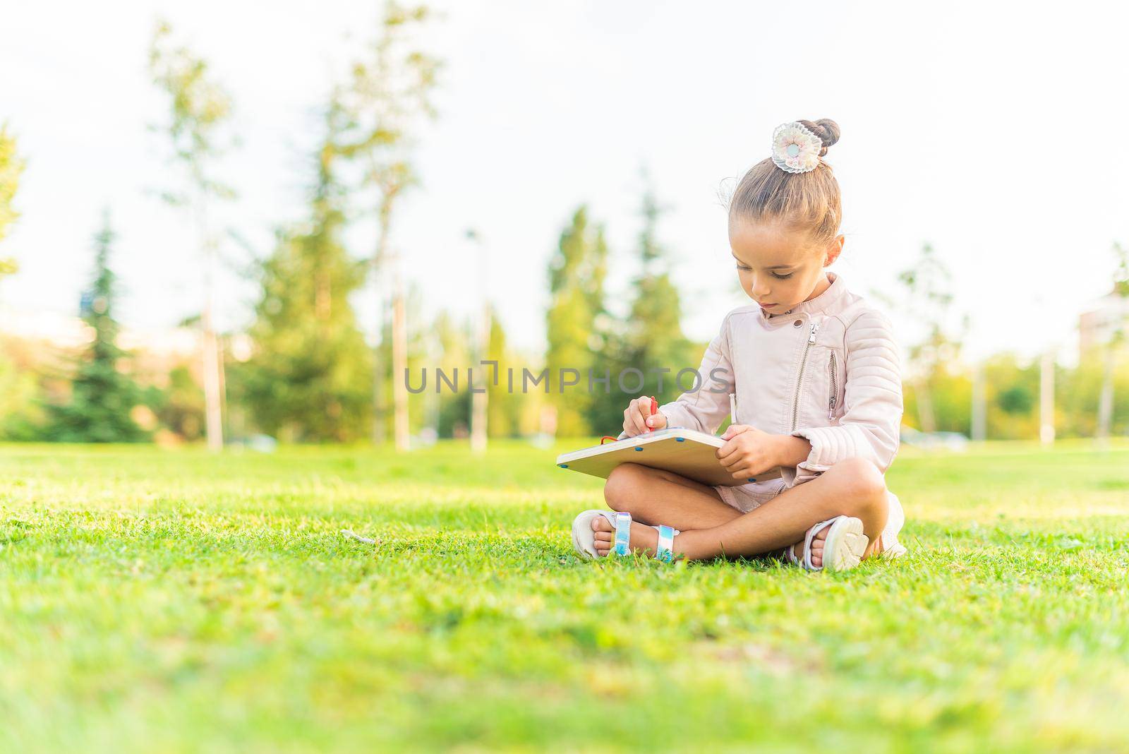 Low angle view of a little girl sitting on the grass in a park playing with a drawing board with copy space. Selective focus.