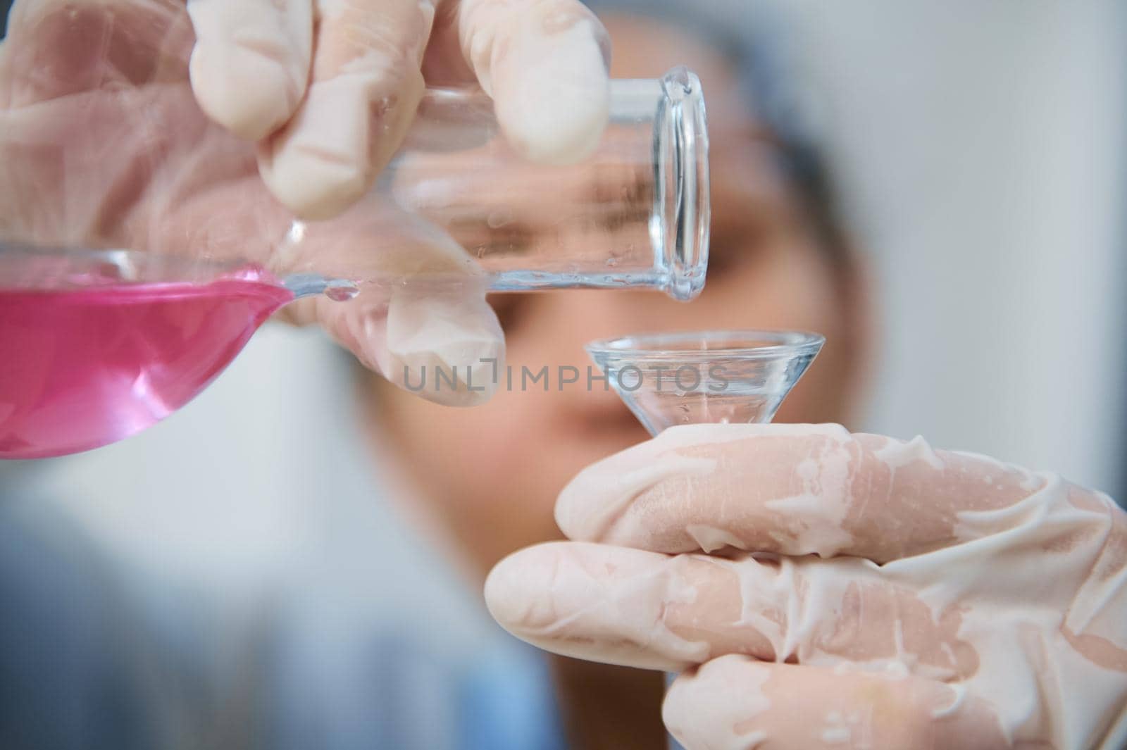 Details: Gloved hands of a blurred laboratory personnel pouring pink liquid chemical substance from a glass flask through a funnel into a test tube while conducting analyzes in a medical laboratory