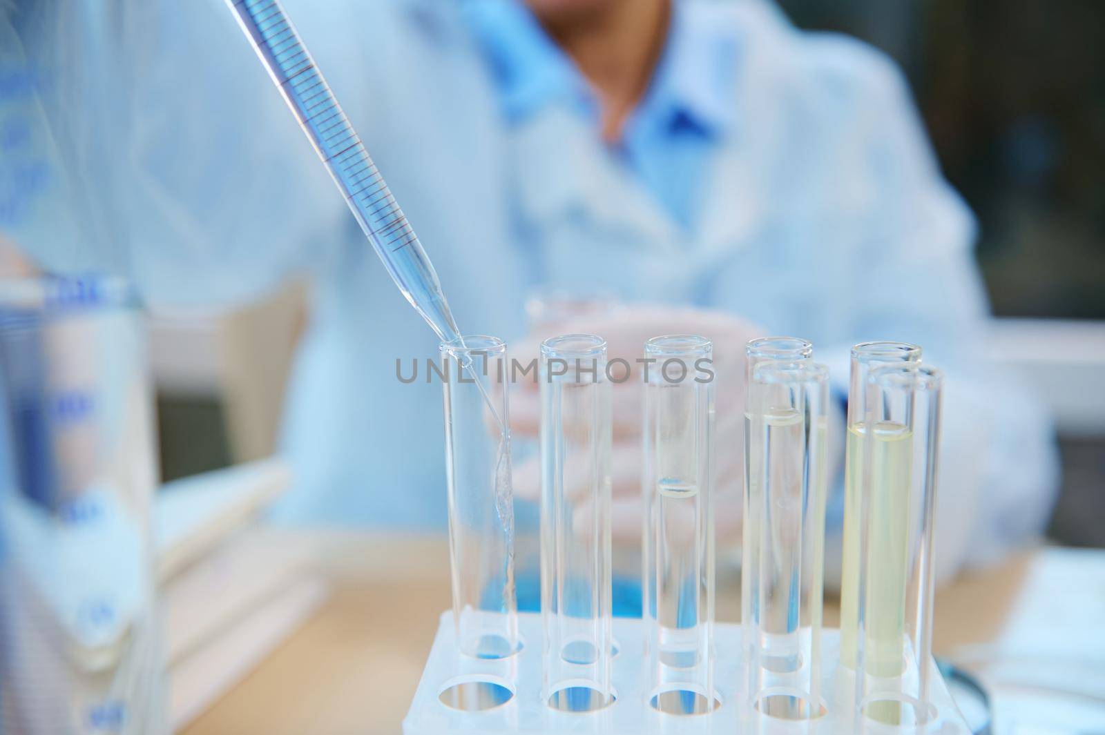 Focus on a glass pipette with a dripping reagent in a test tube on tripod, over blurred background of a chemical laboratory assistant conducting clinical analysis in the biological chemical laboratory