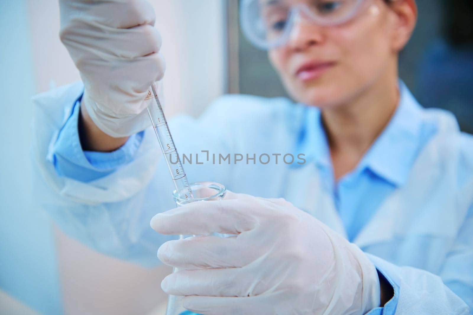 Focus on glass laboratory pipette in gloved hands of blurred female pharmacologist, medical biologist, scientist chemist dripping few drops of chemical reagent into flask while conducting experiment