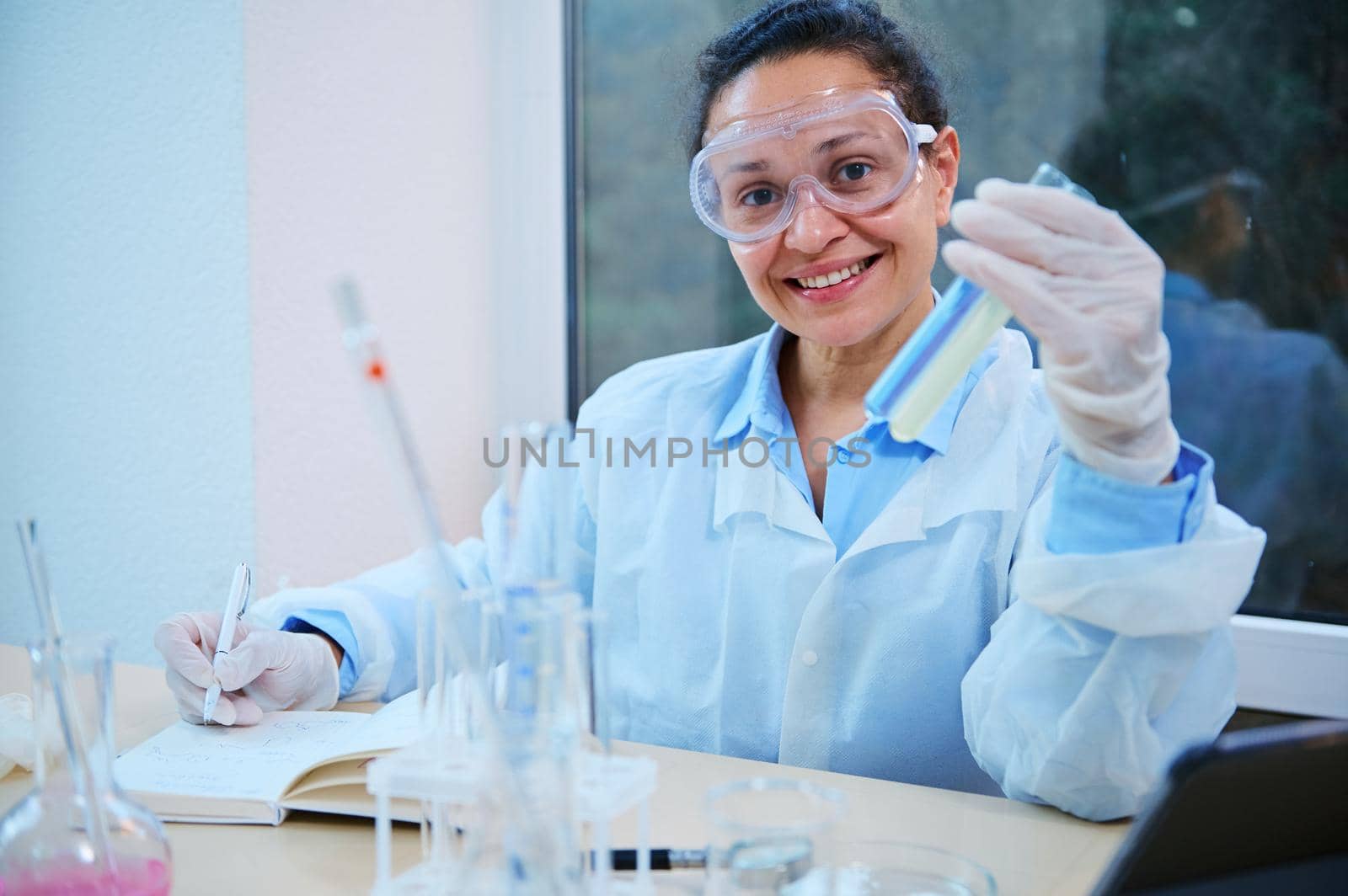 Positive multi-ethnic woman, scientist, medical researcher in safety labwear, holds test tubes with chemical substances, makes notes on nopetapd and smiles looking at camera, in a science laboratory