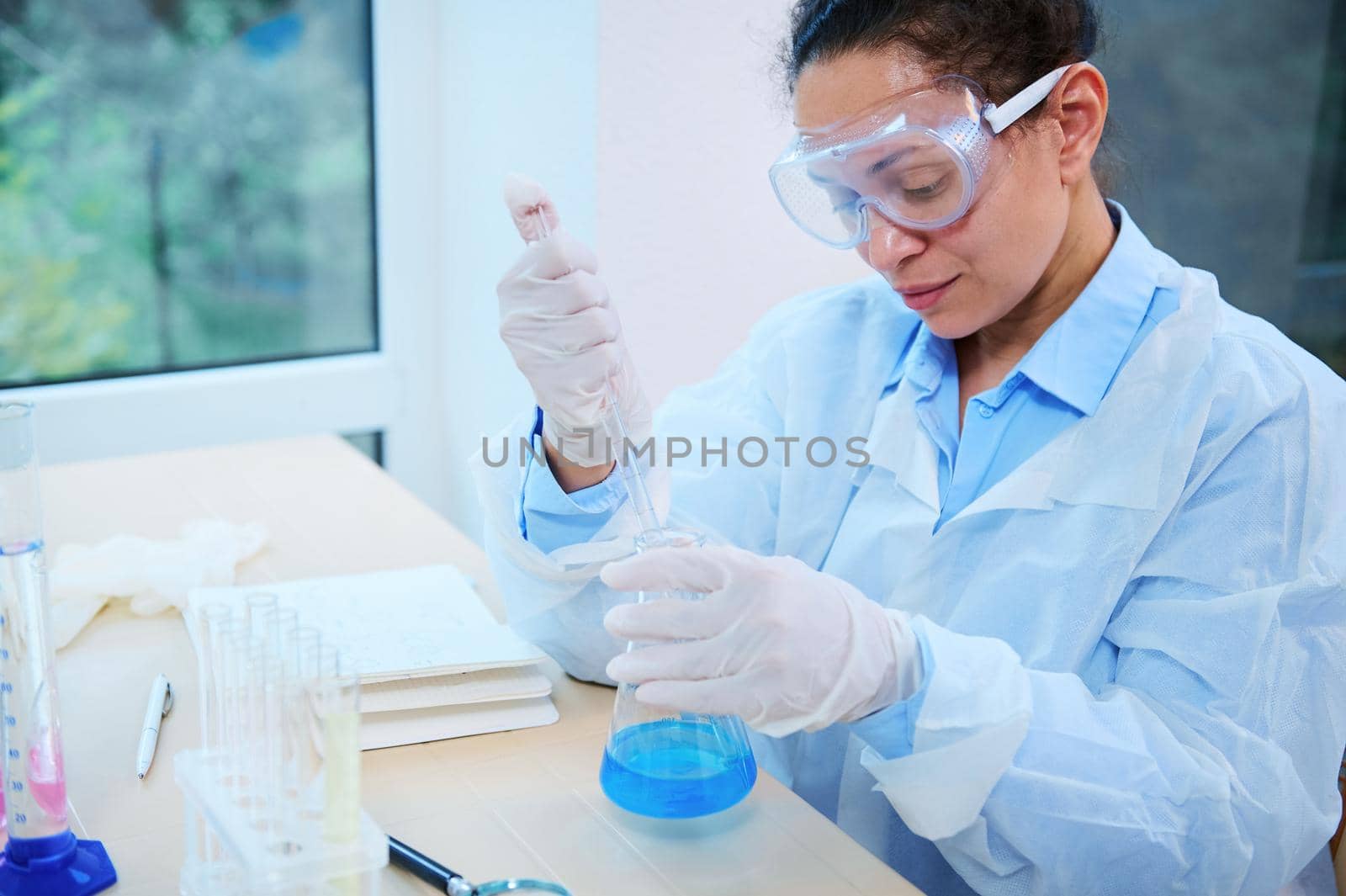 African American woman, scientist pharmacologist pipetting the blue chemical liquid in a flat-bottomed flask, sitting at a table in a light laboratory interior, conducting a science experiment