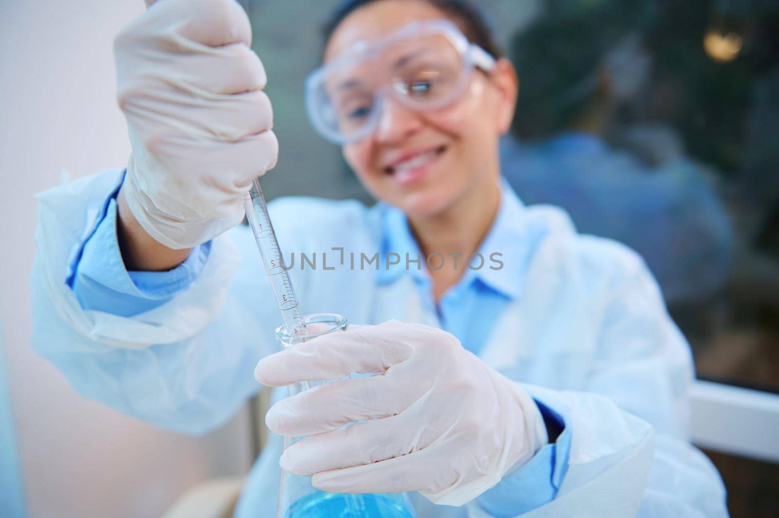 Selective focus on glass dropper in gloved hands of blurred woman scientist chemist wearing safety goggles and lab coat, doing science experiments, pipetting chemical solution in biological laboratory